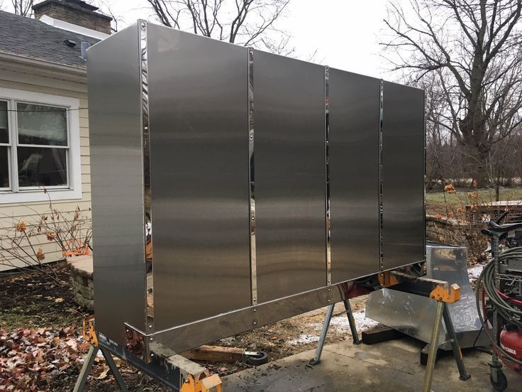 Large stainless steel enclosure beside a house in a yard, with tools and a trailer nearby