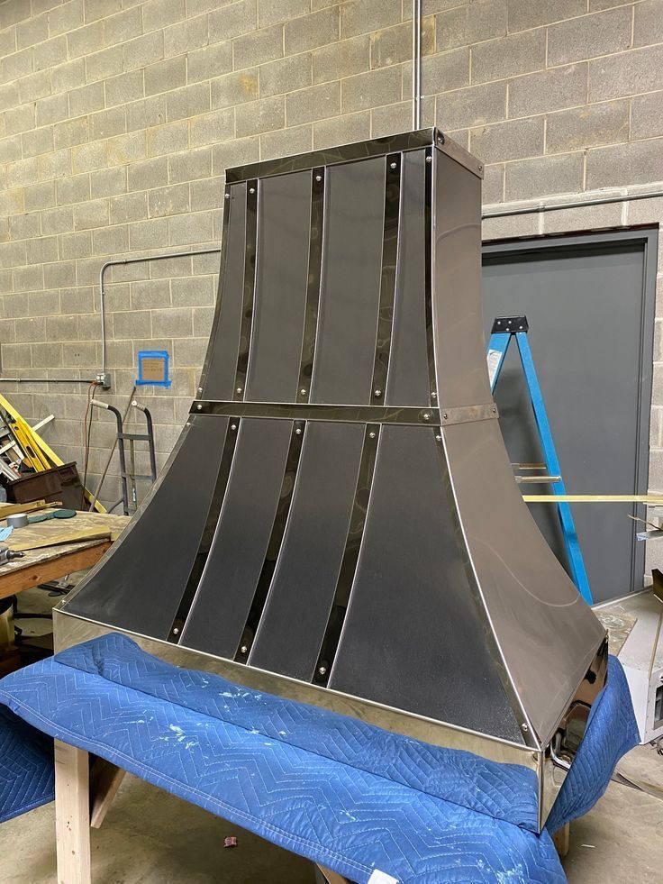 Large black metal duct or chute on a blue-wrapped worktable in a workshop, against a cinderblock wall.