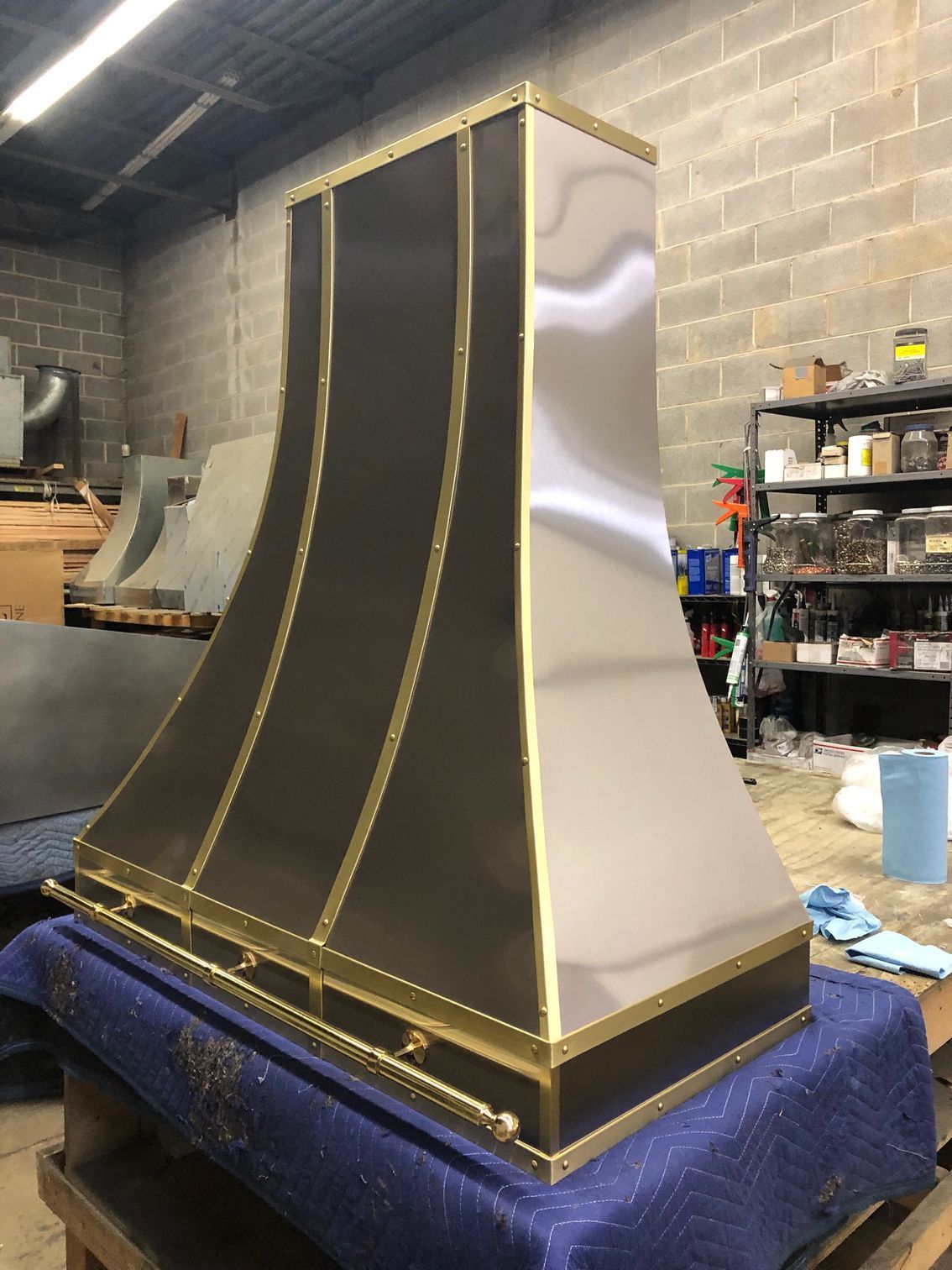 Large black-and-gold metal hood on a blue padded stand in a workshop, with shelves and tools in the background
