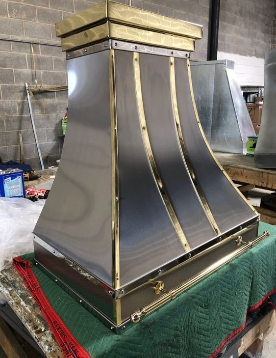 Shiny stainless-steel range hood with gold trim, shown on a workbench in a workshop.