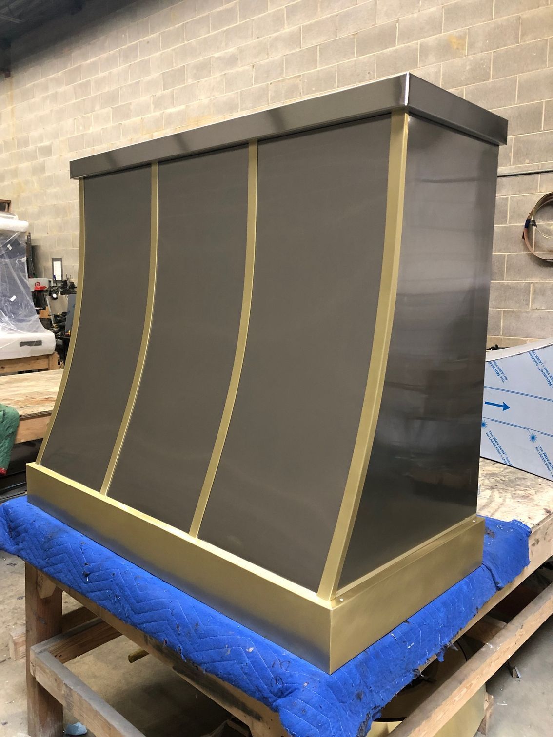 Large gray metal hood with gold vertical trim on a blue-wrapped pallet in a workshop