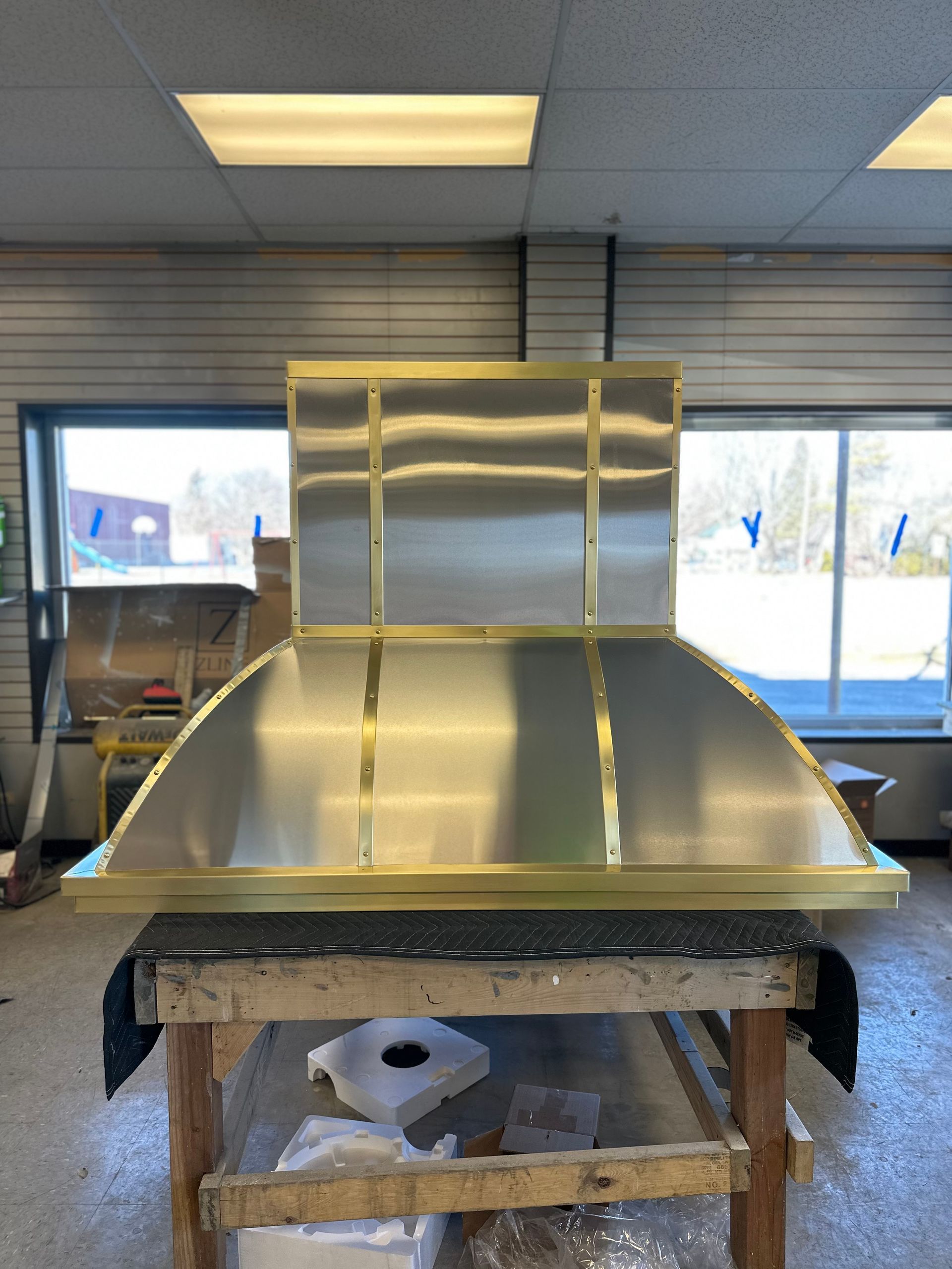 A metal hood or duct on a workbench in a workshop, with windows and tools in the background.