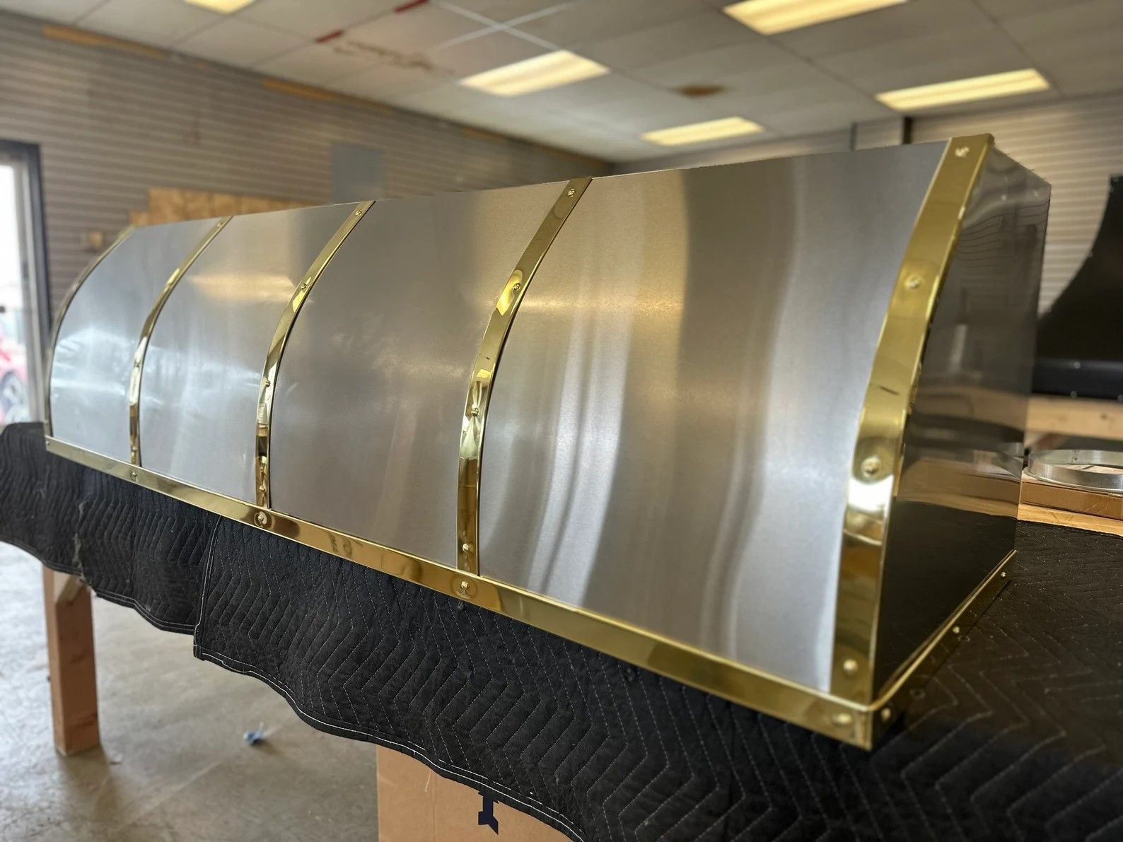 Large stainless steel industrial hood or duct section with gold trim on a stand in a workshop