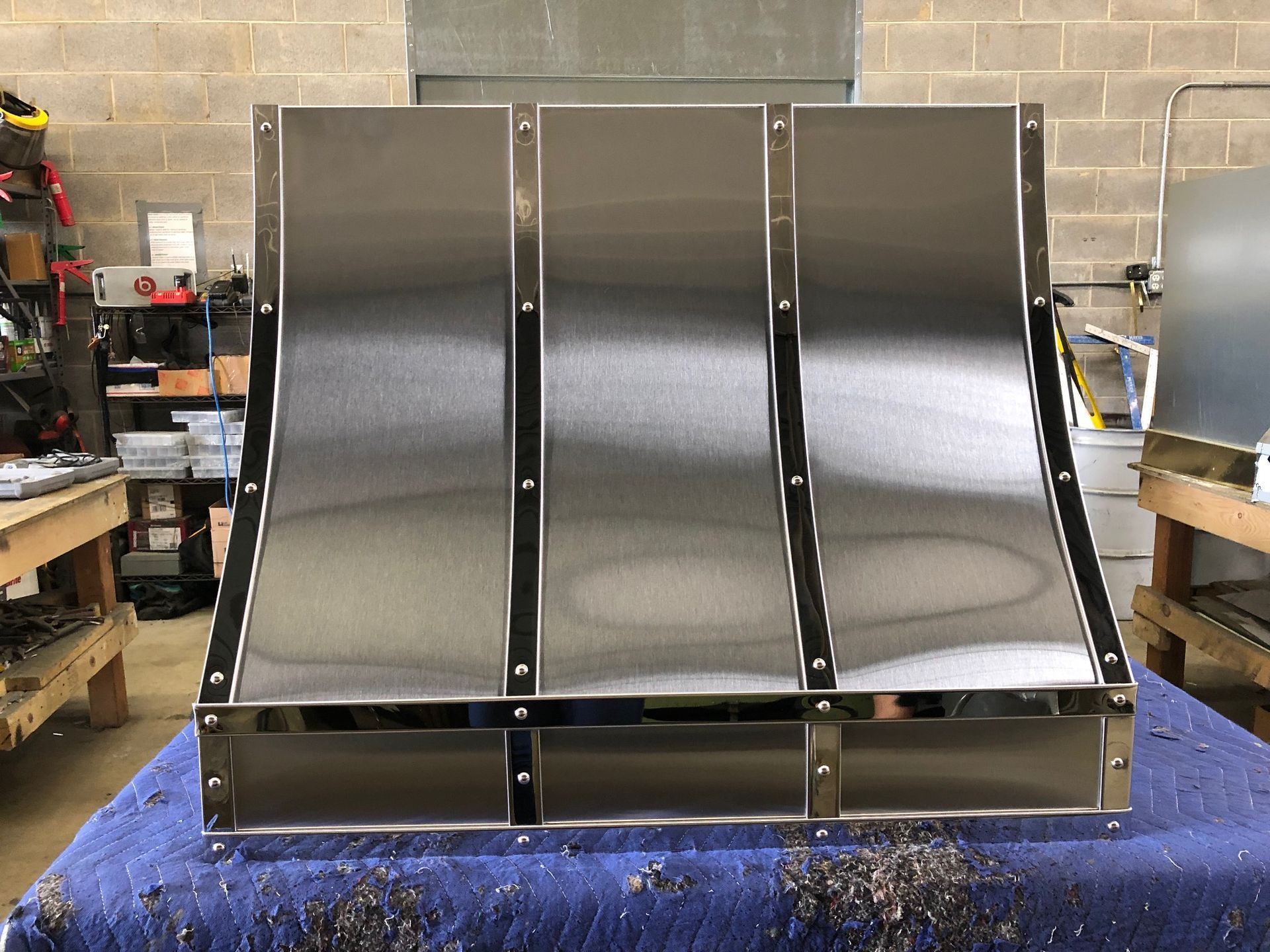 Black metal machine attachment with three curved panels on a blue table in a workshop