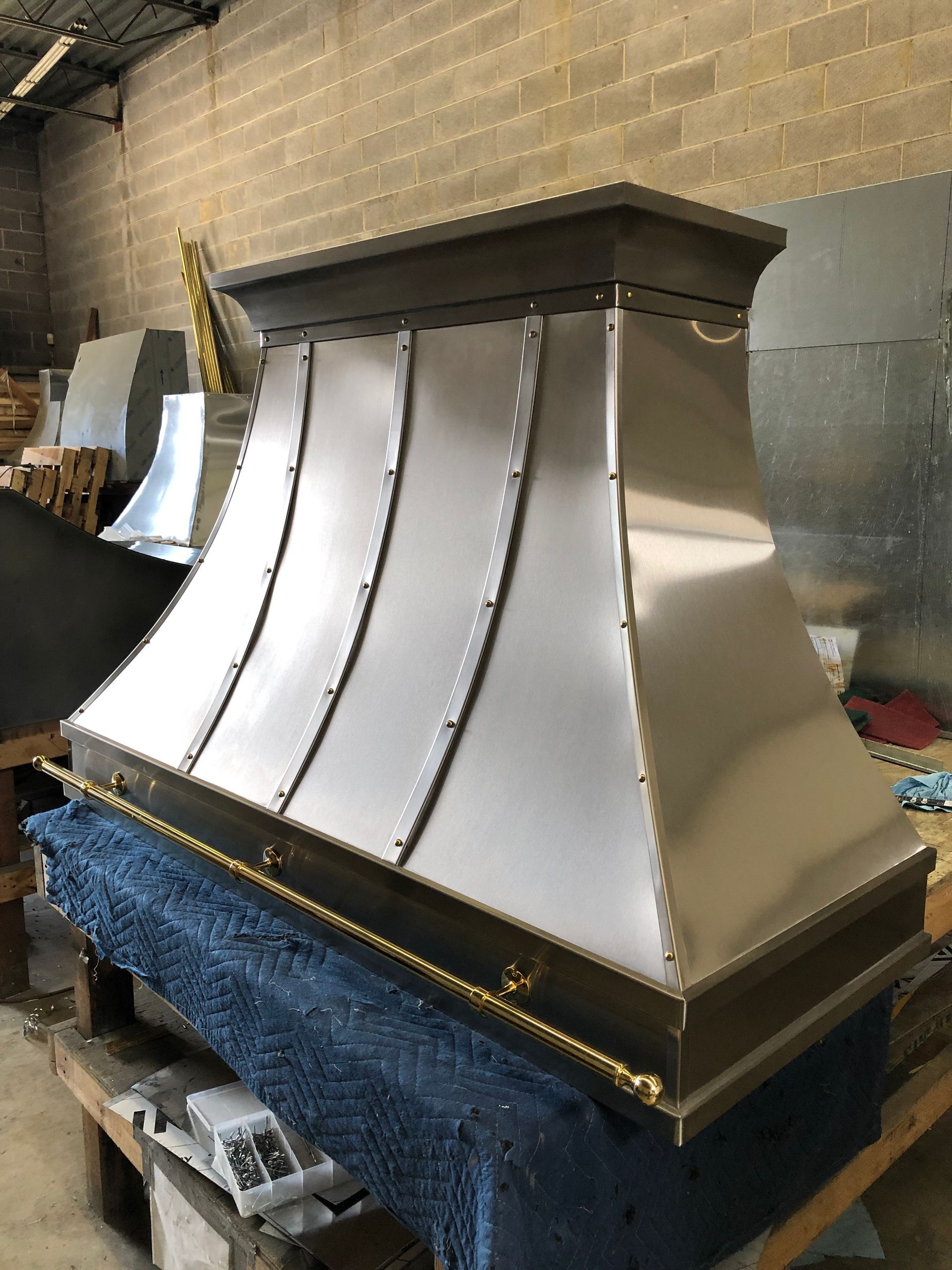 Stainless steel range hood on a workbench in a workshop, with curved panels and a boxed top.