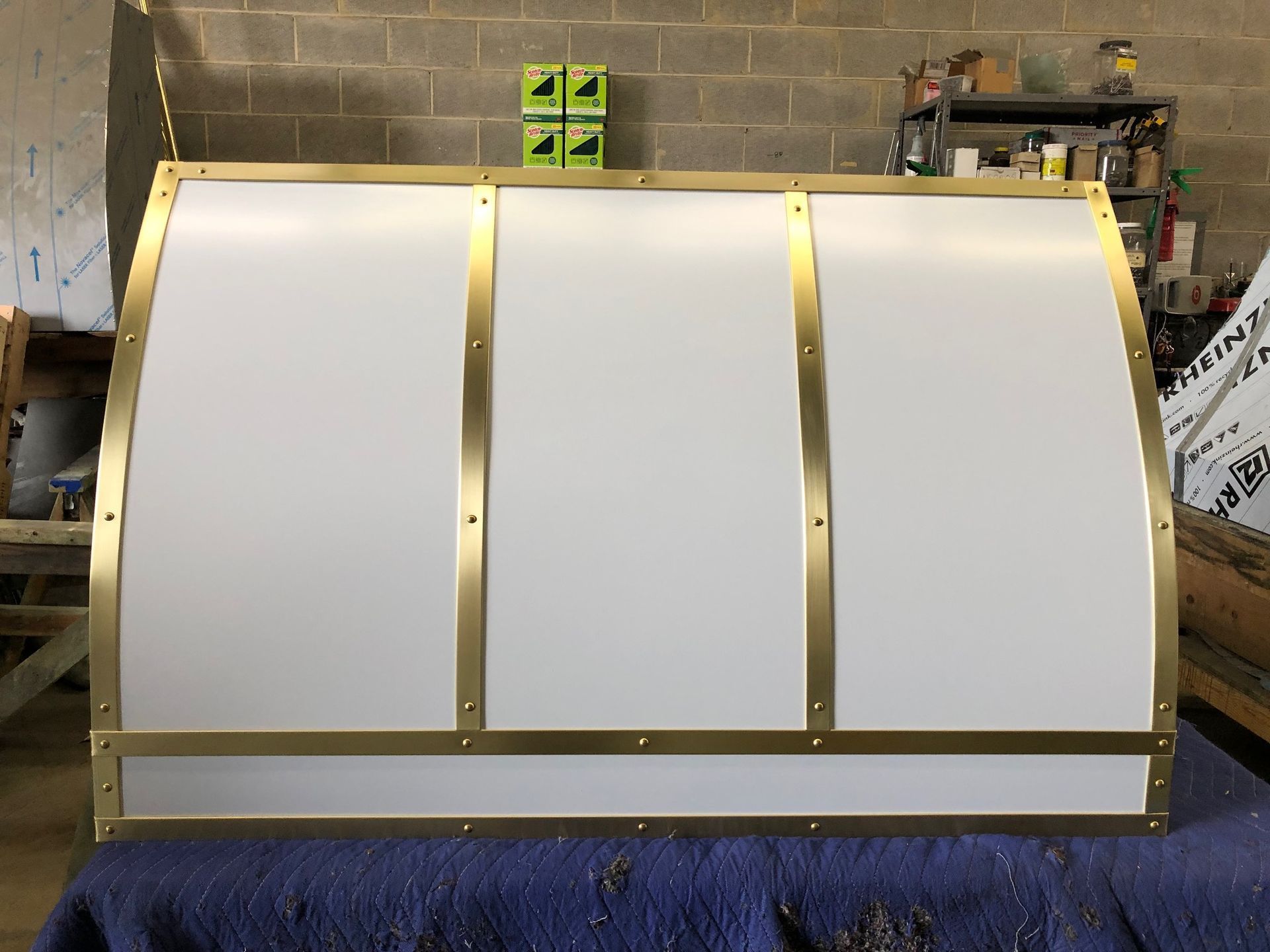 White curved panel with brass trim standing in a workshop, partially wrapped in blue material