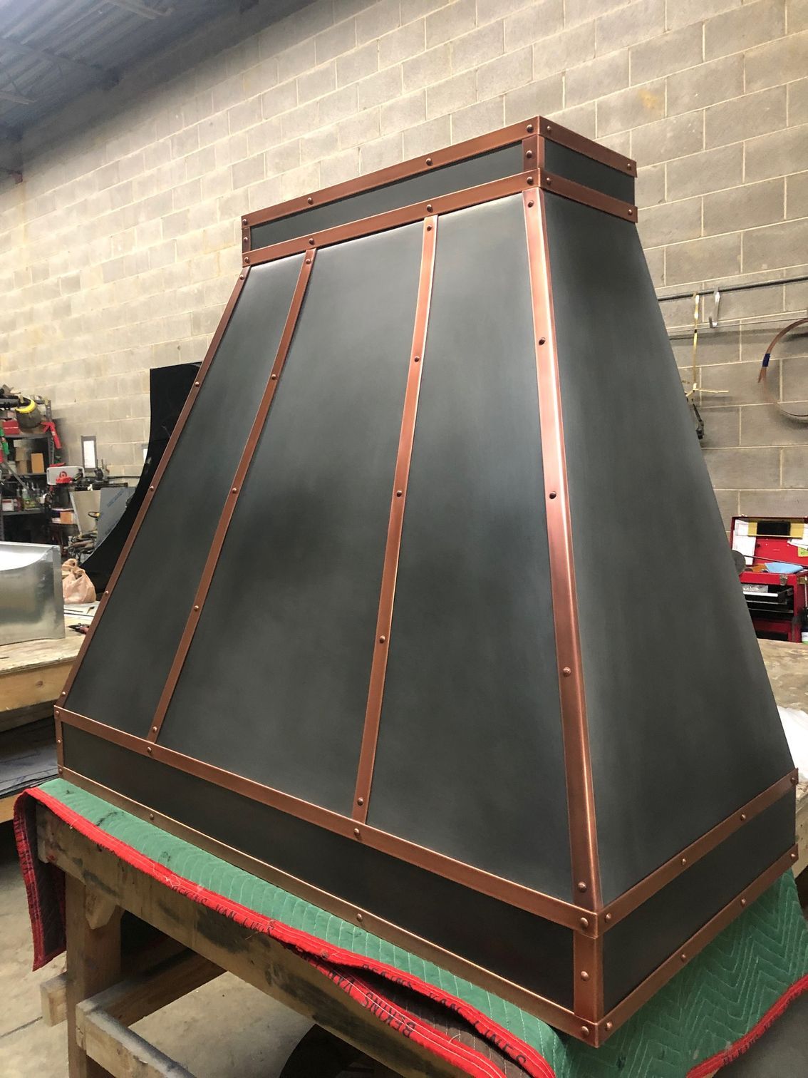 Large black-and-copper range hood in a workshop, propped on a green-covered table.
