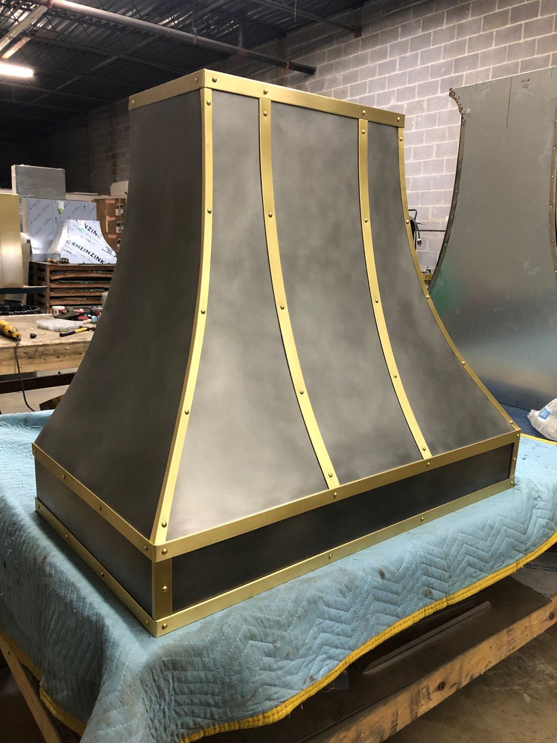 Metal industrial hood prototype with gold trim on a blue padded workbench in a workshop