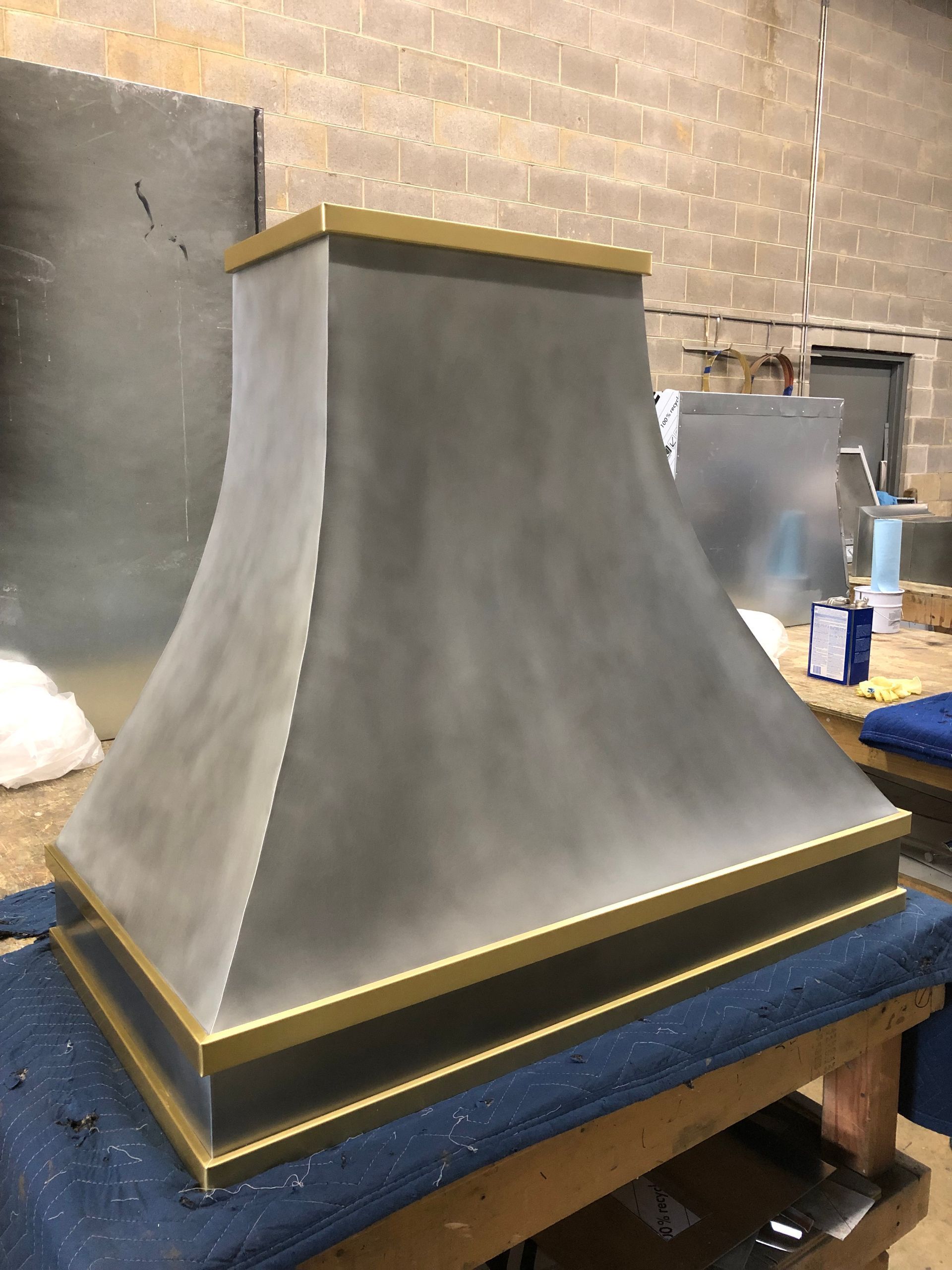 Large unfinished gray metal range hood with gold trim on a workbench in a workshop