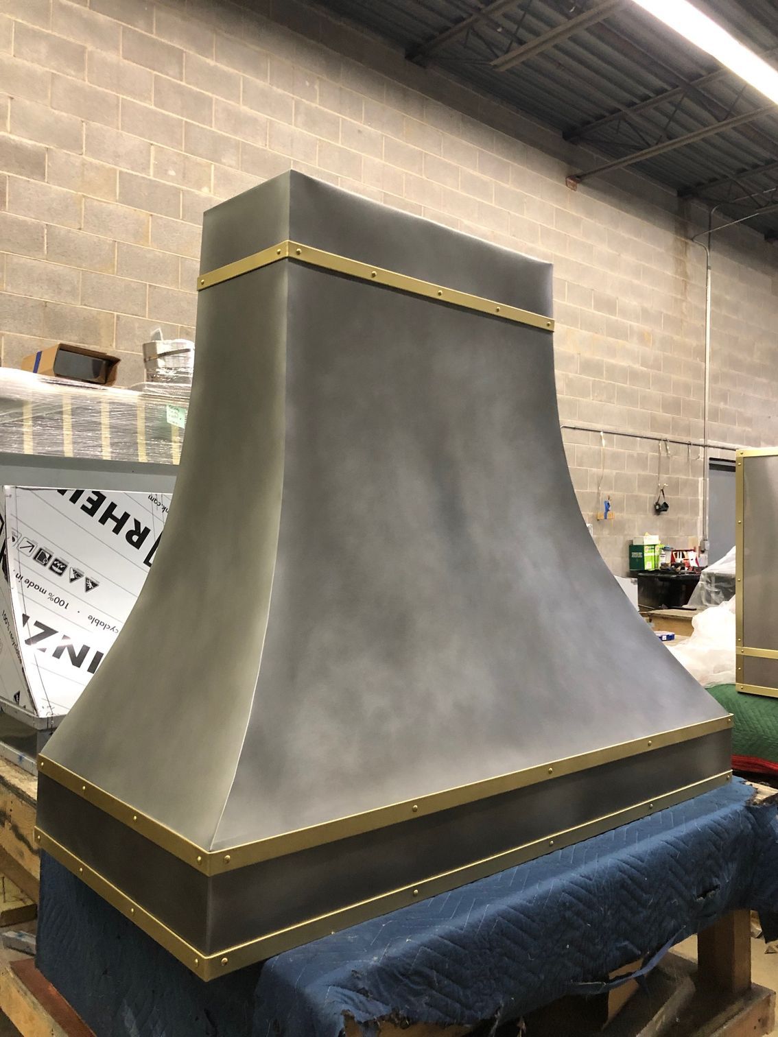 Large gray metal hood with gold trim sitting on a blue-draped base in a workshop.