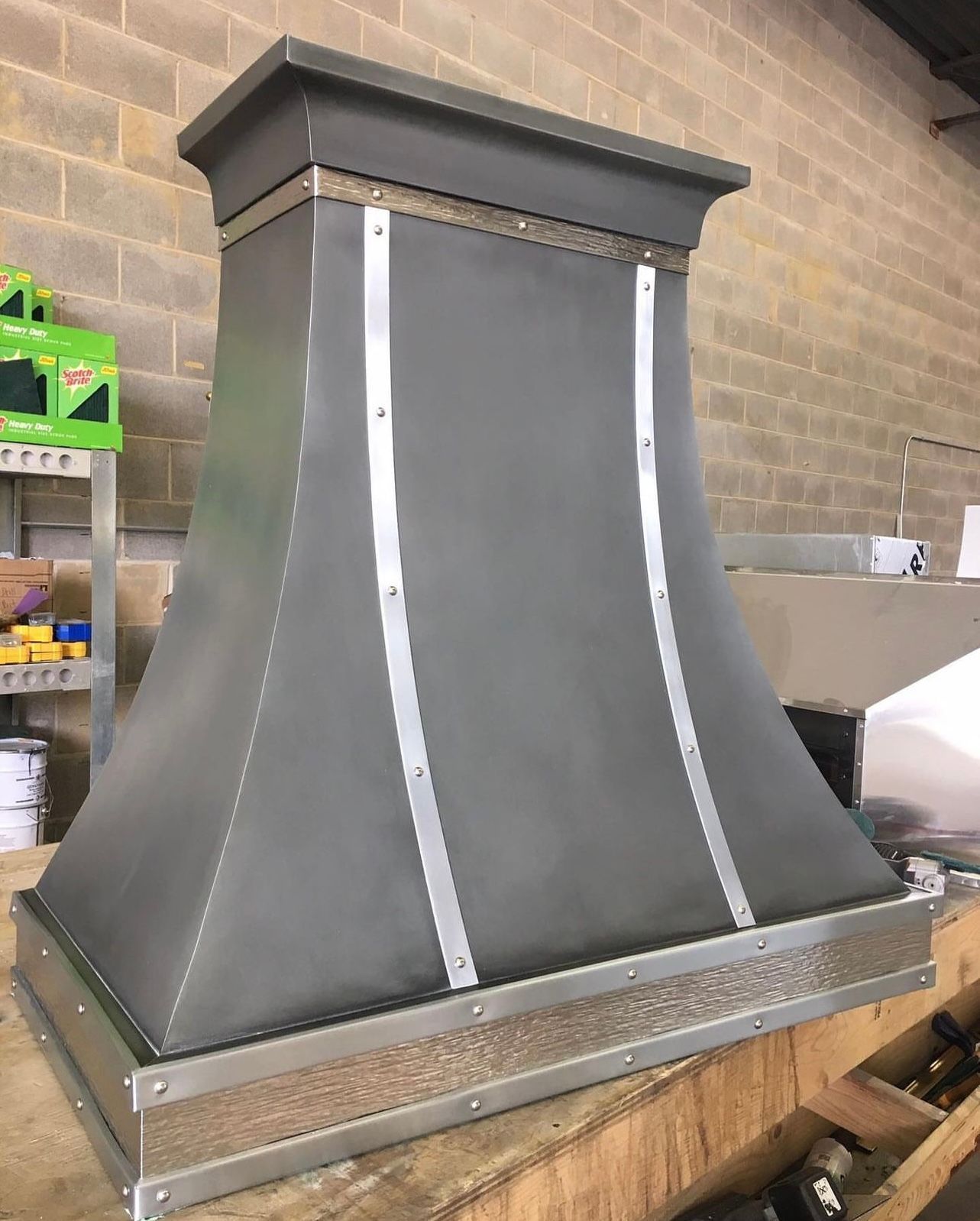 Black metal vent hood with silver trim in a workshop, viewed from the front
