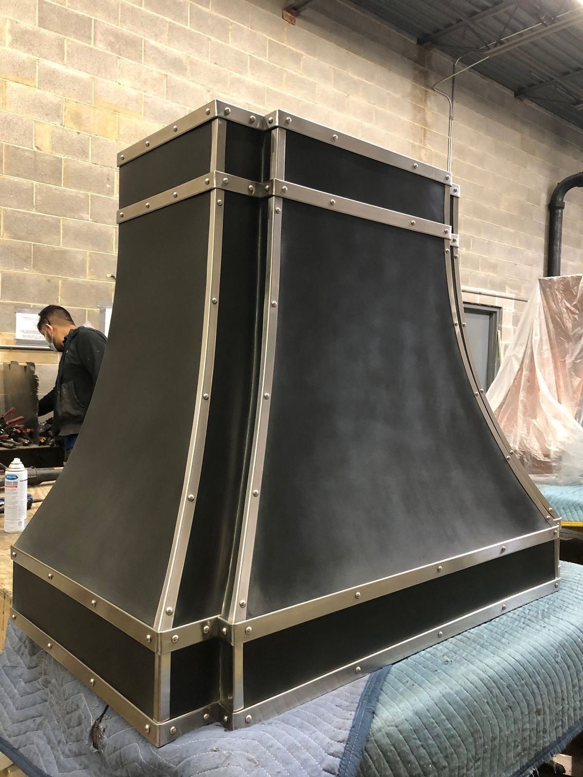 Large black industrial metal hood or duct on a workbench in a workshop, with brick walls in the background