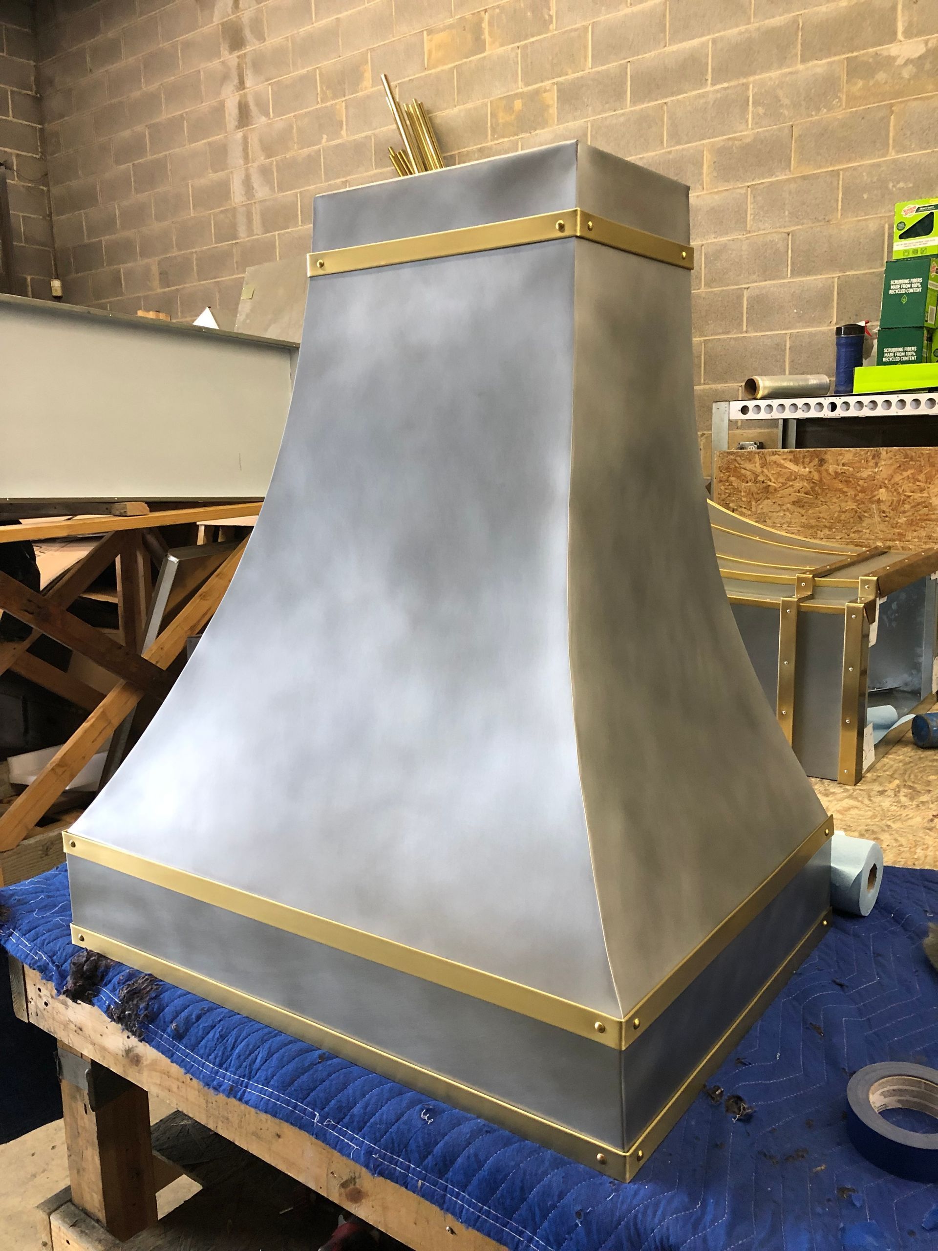 Metallic gray stove hood with gold trim on a blue padded base in a workshop or studio.