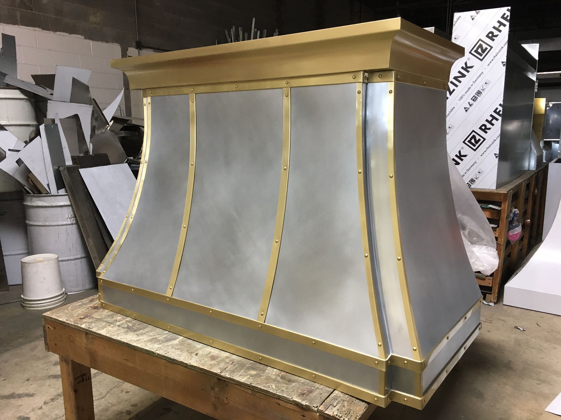 Metal hood or vent cover on a wooden workbench in a workshop, with silver panels and gold trim.