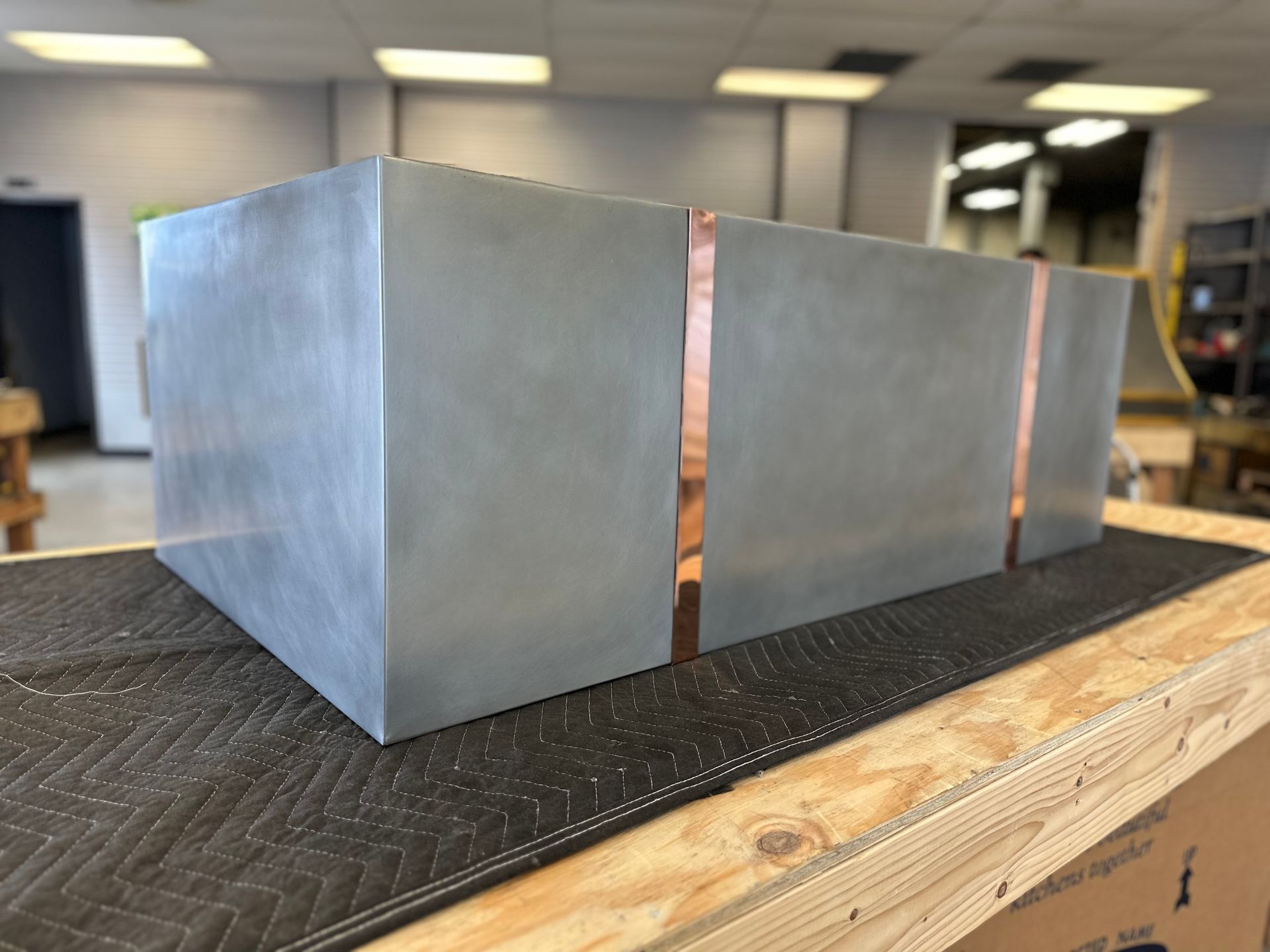 Metal box-like structure with narrow vertical copper-colored seams on a workbench in a workshop