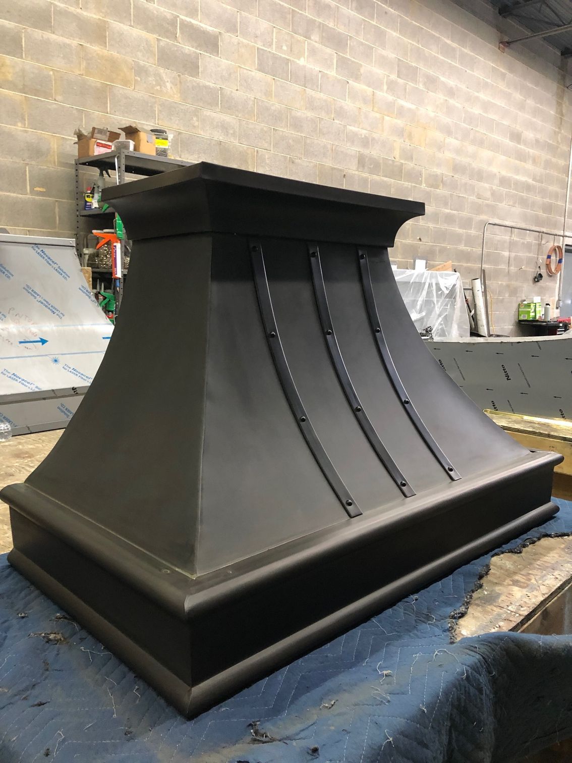 Black metal vent hood with decorative ribbed front, displayed on a worktable in a workshop.