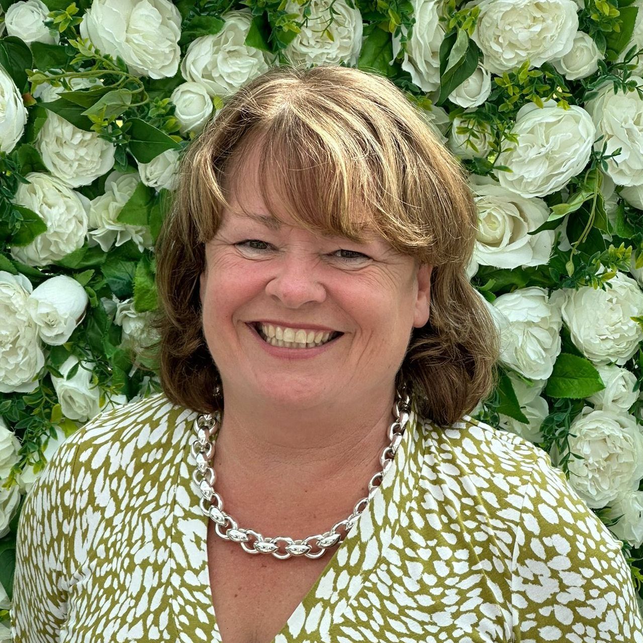 A woman in a yellow jacket is smiling in front of a wall of white flowers