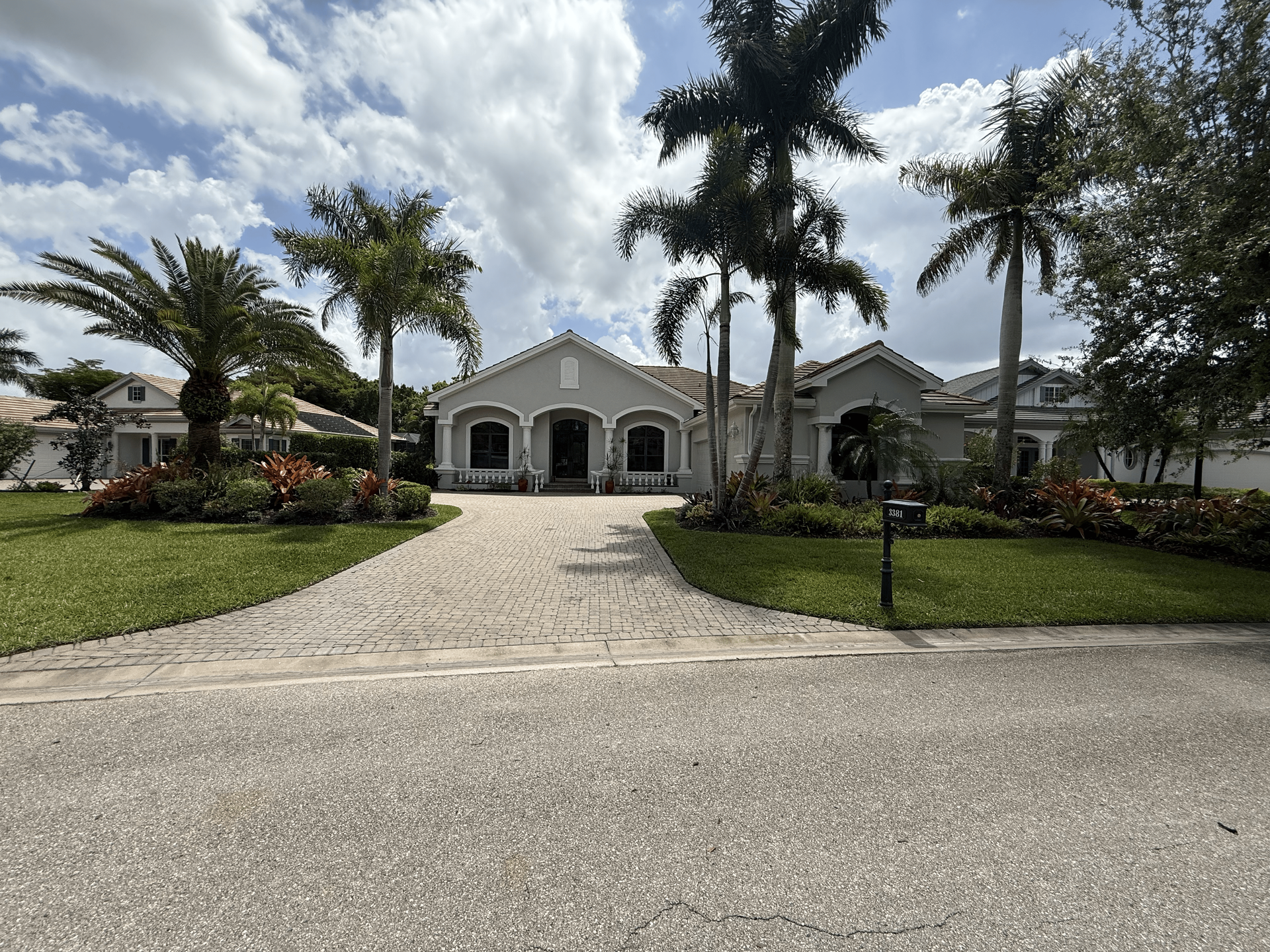 Large white house with arched porch, gravel driveway, palm trees, and landscaped yard under a cloudy sky