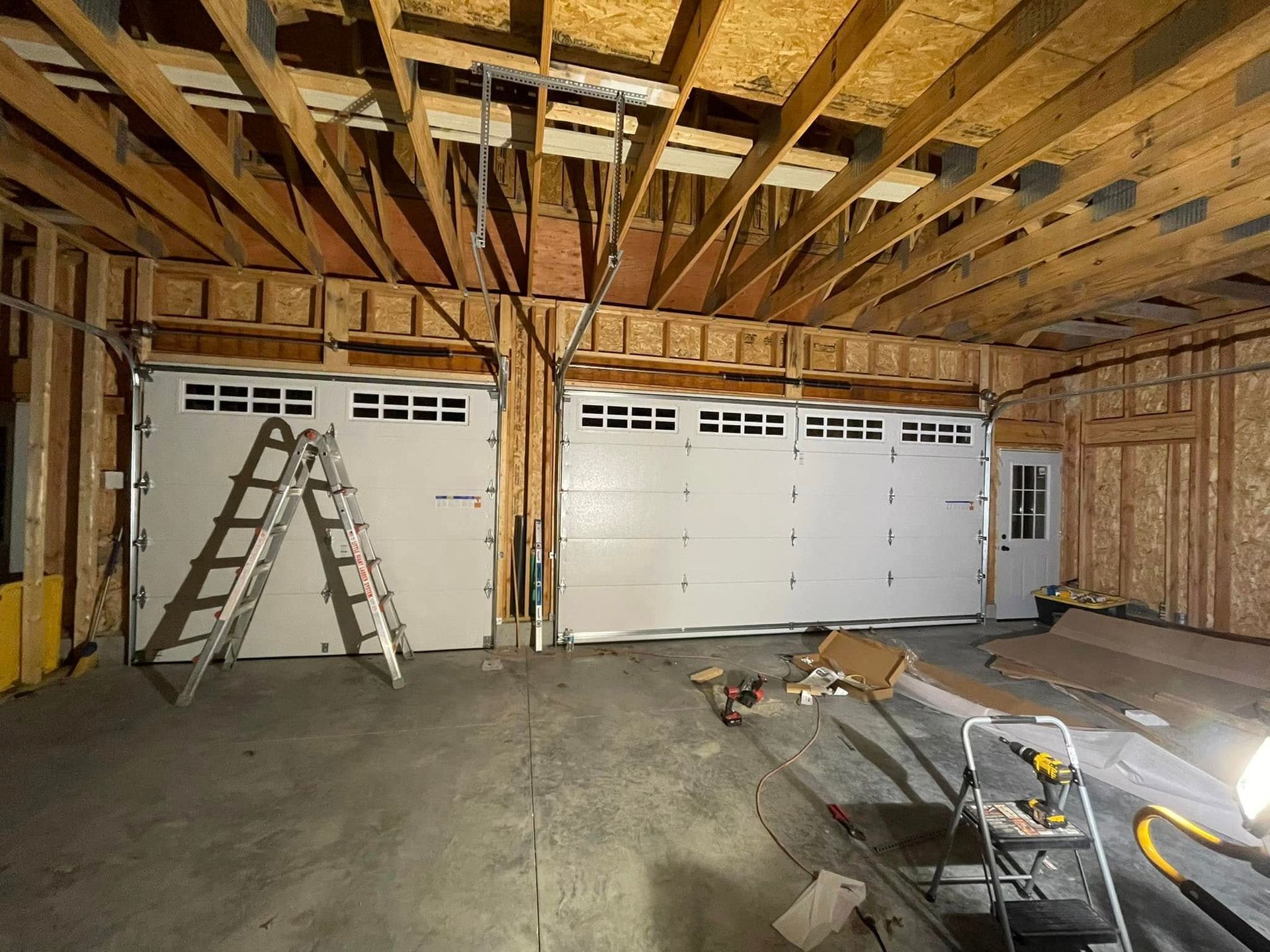 Unfinished garage interior with exposed rafters, two ladders, and a large white garage door under construction