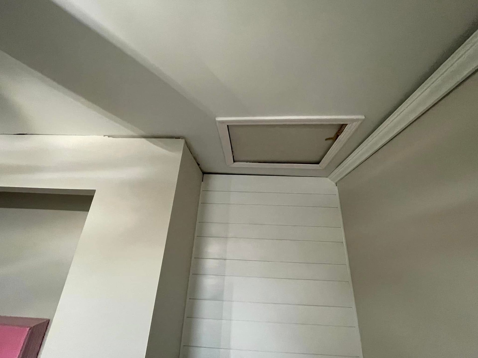 Corner of a room with white walls and a small ceiling hatch or attic access panel.