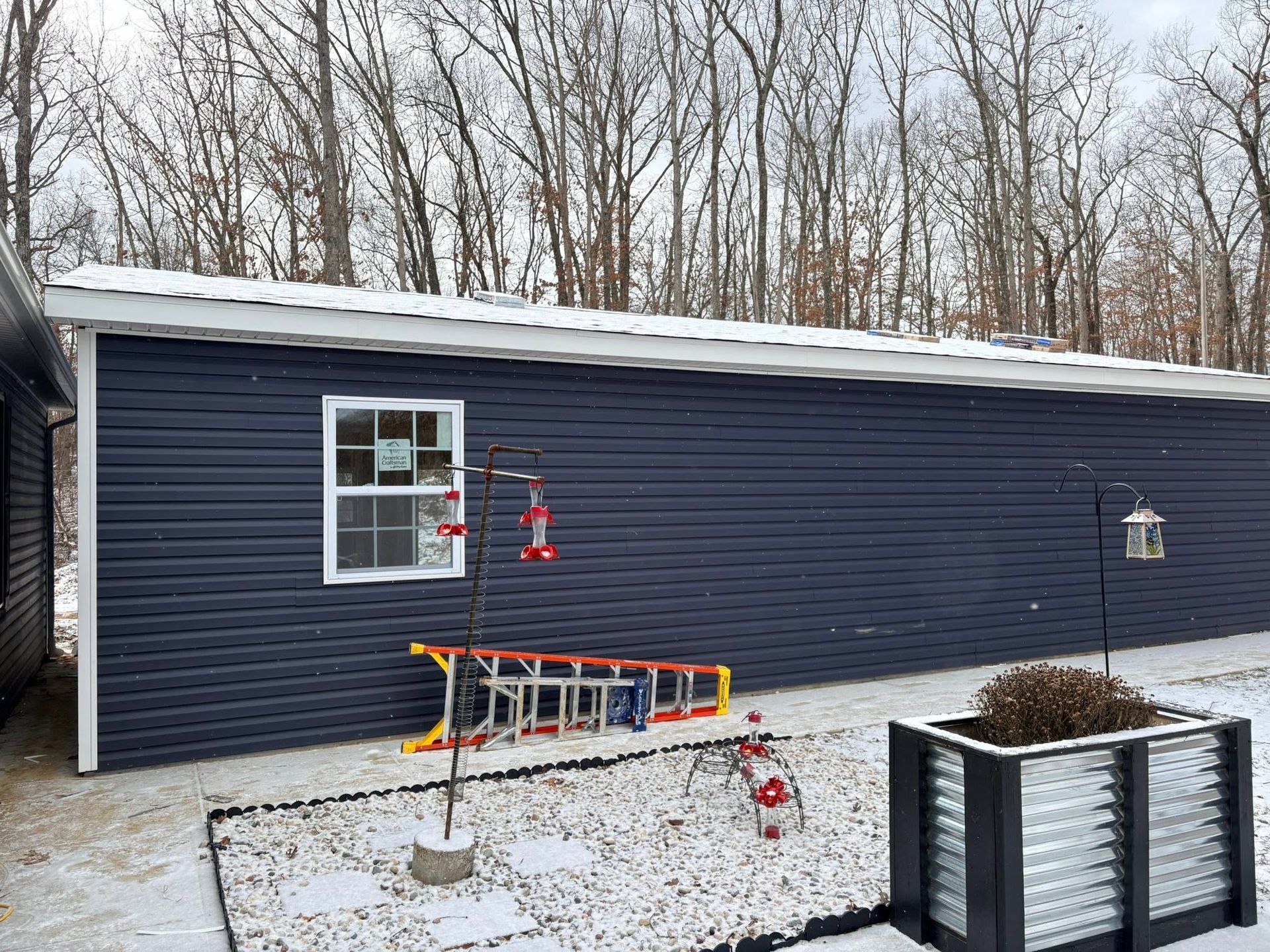 Snow-dusted dark blue garage with a window, ladder, and planter in a leafless yard