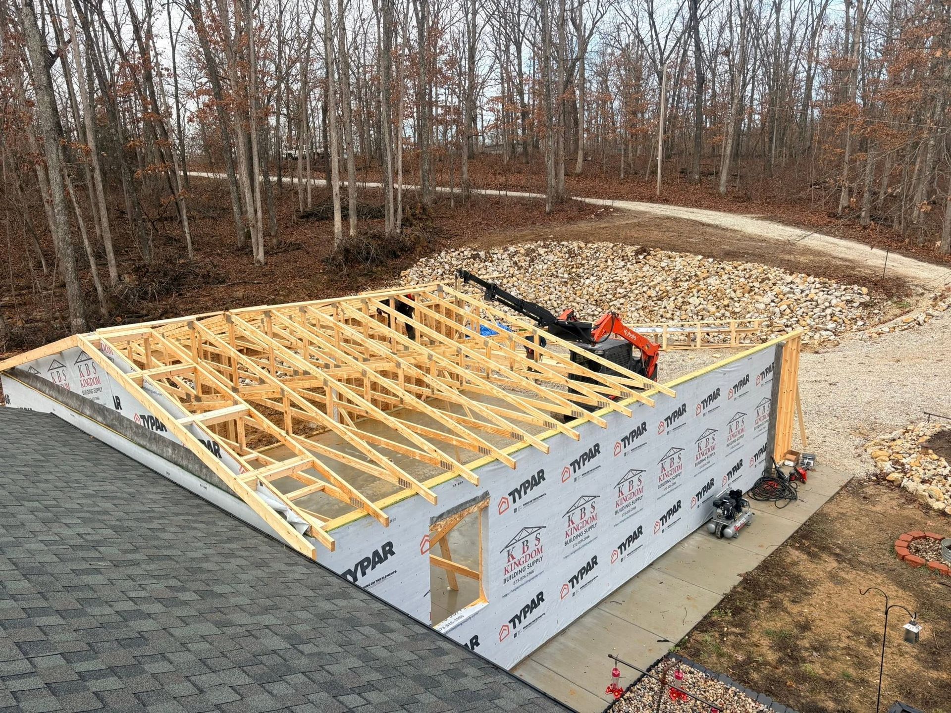 House foundation under construction with wooden roof framing and white wall panels beside a wooded lot