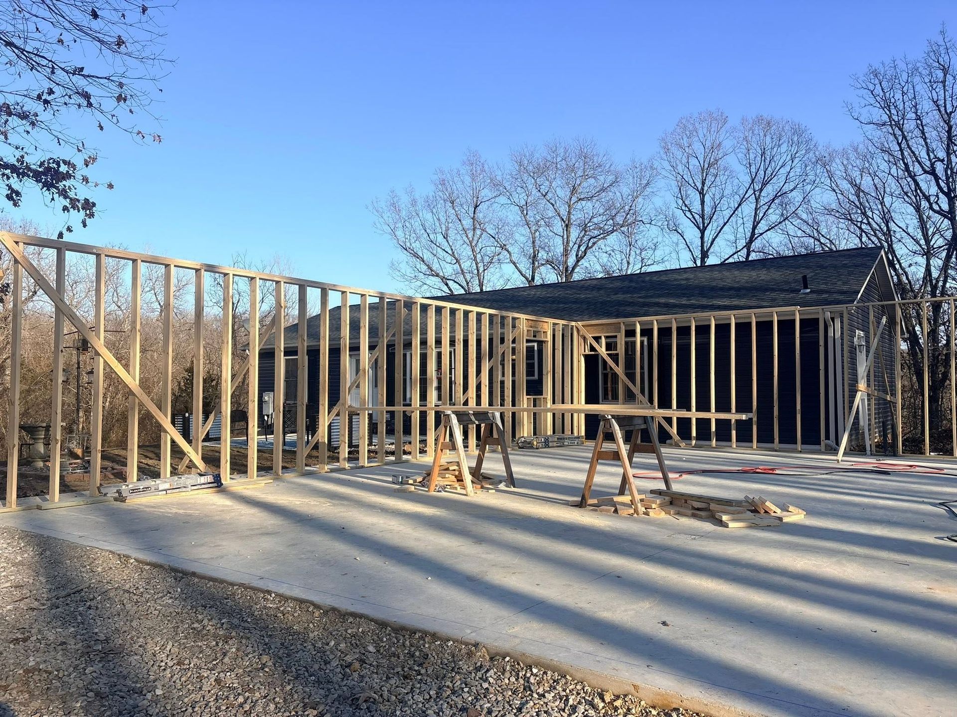 Framed house walls under construction on a concrete foundation with two sawhorses in front and trees behind