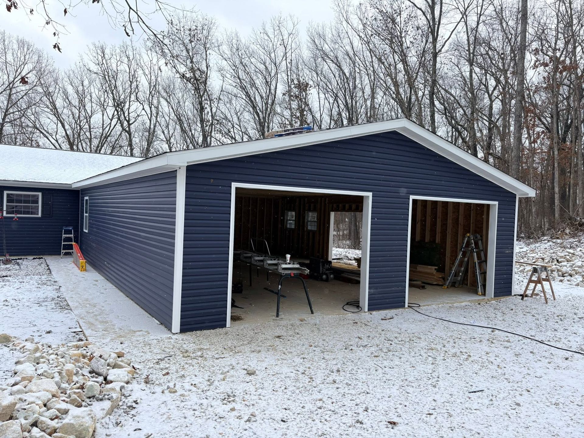 Snow-covered blue garage with open bays in a wooded yard