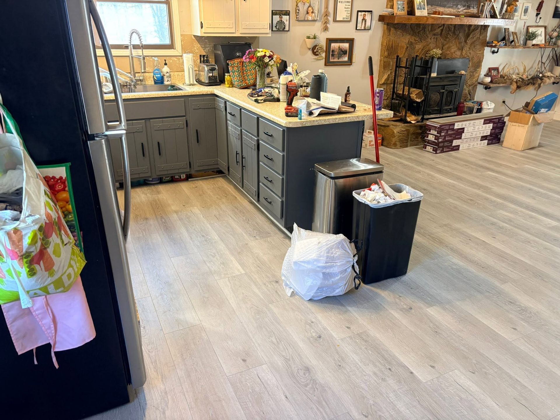 Kitchen with gray cabinets, light wood floor, trash bins, and a fireplace in the background.