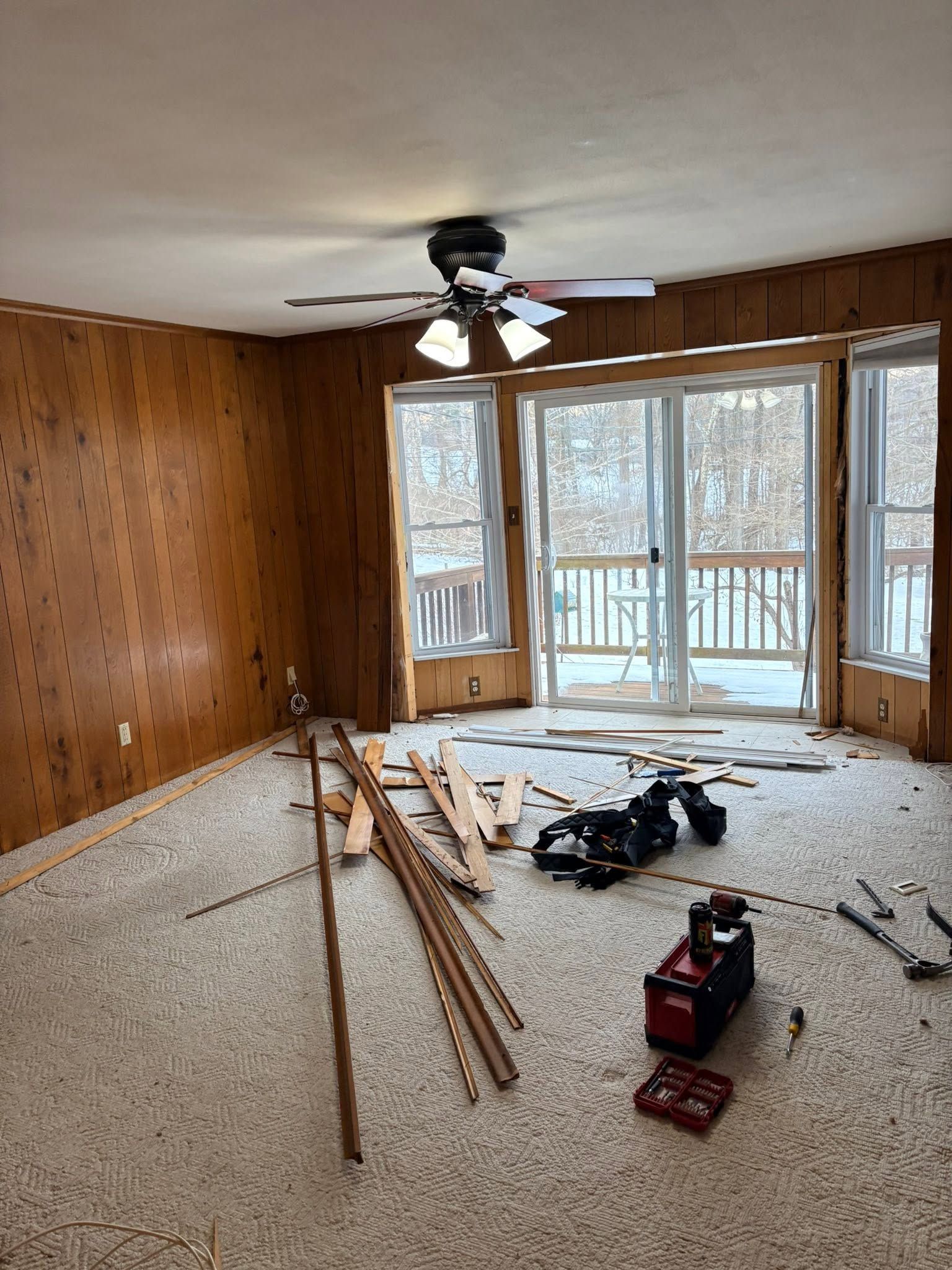 Wood-paneled room under renovation with tools, wood planks, and a ceiling fan near large windows.
