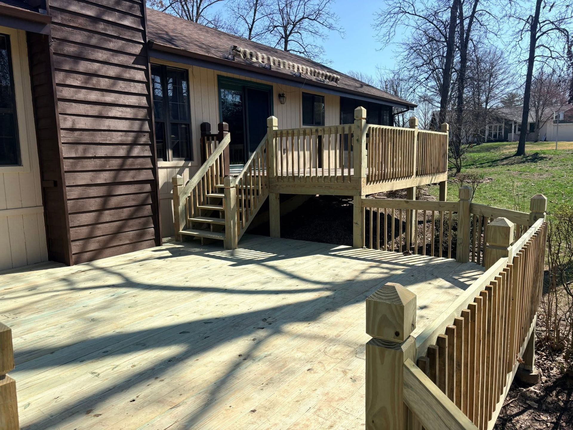 Wooden deck and stairs beside a house, with a fenced patio and yard in sunlight.