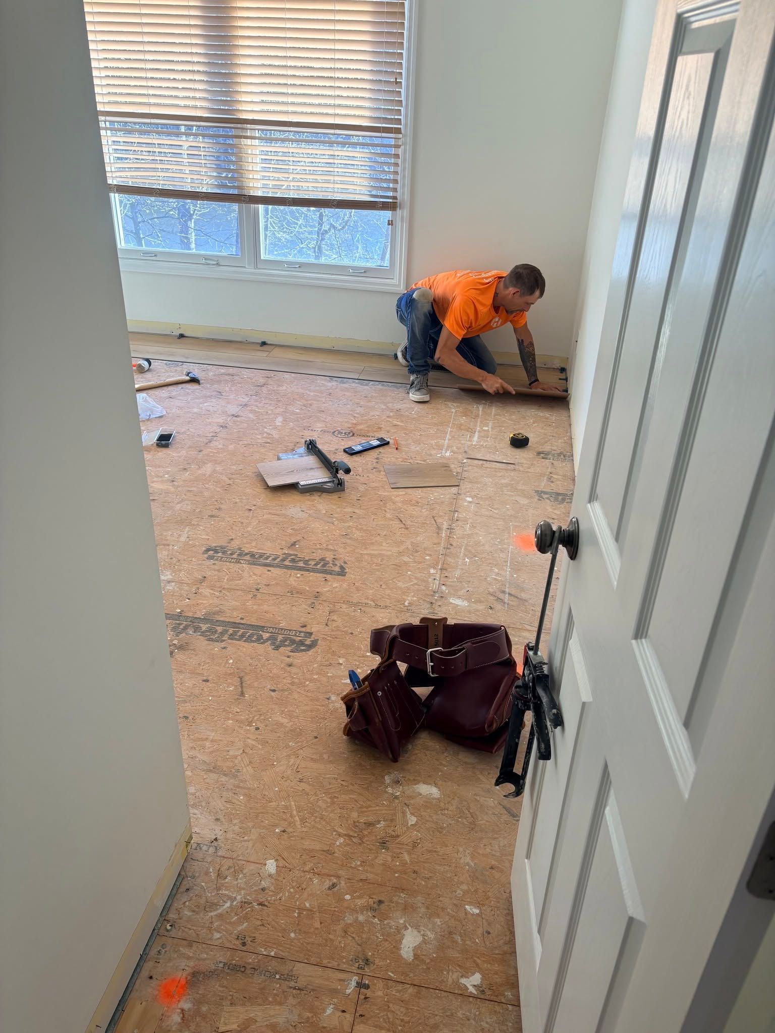 Worker kneeling on a carpeted floor, installing trim in an empty room with tools and debris.