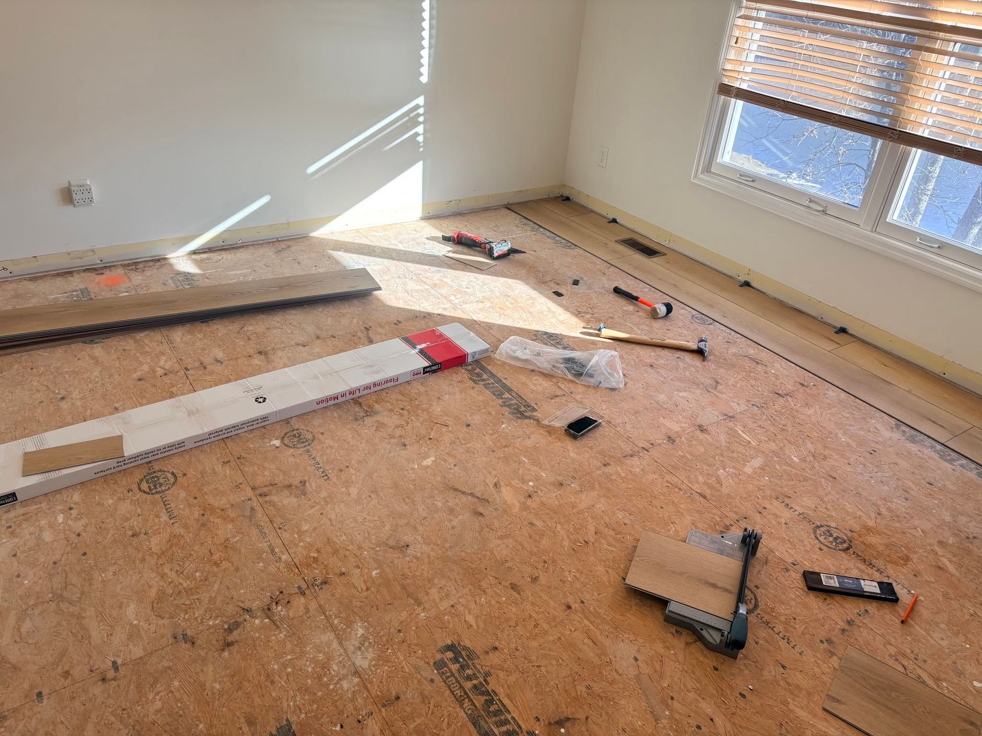 Empty room with unfinished subfloor, scattered tools, and sunlight through windows.
