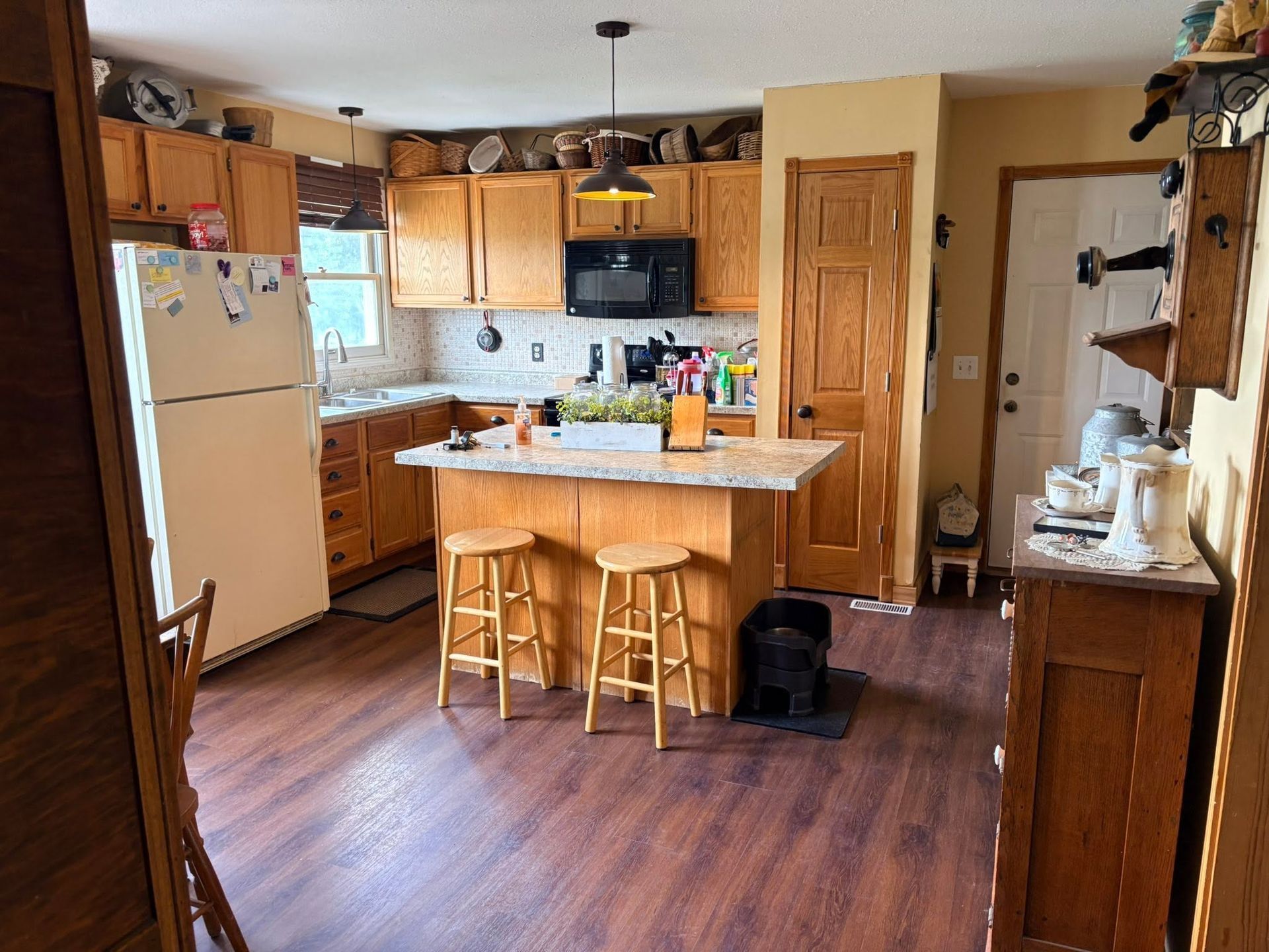 Warm kitchen with wooden cabinets, central island, two stools, and hardwood floor; fridge and sink visible.
