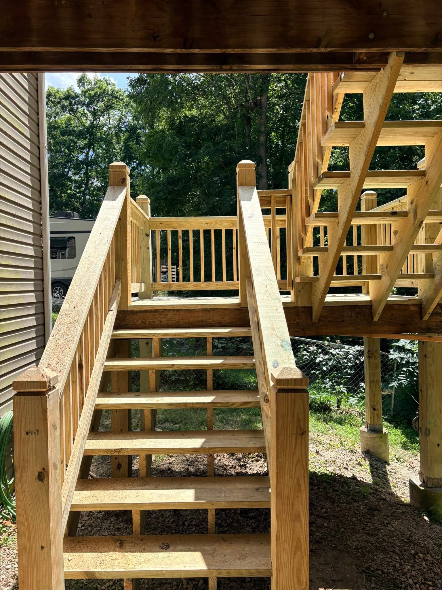 Wooden outdoor stairs under a deck, leading up beside a house with trees in the background