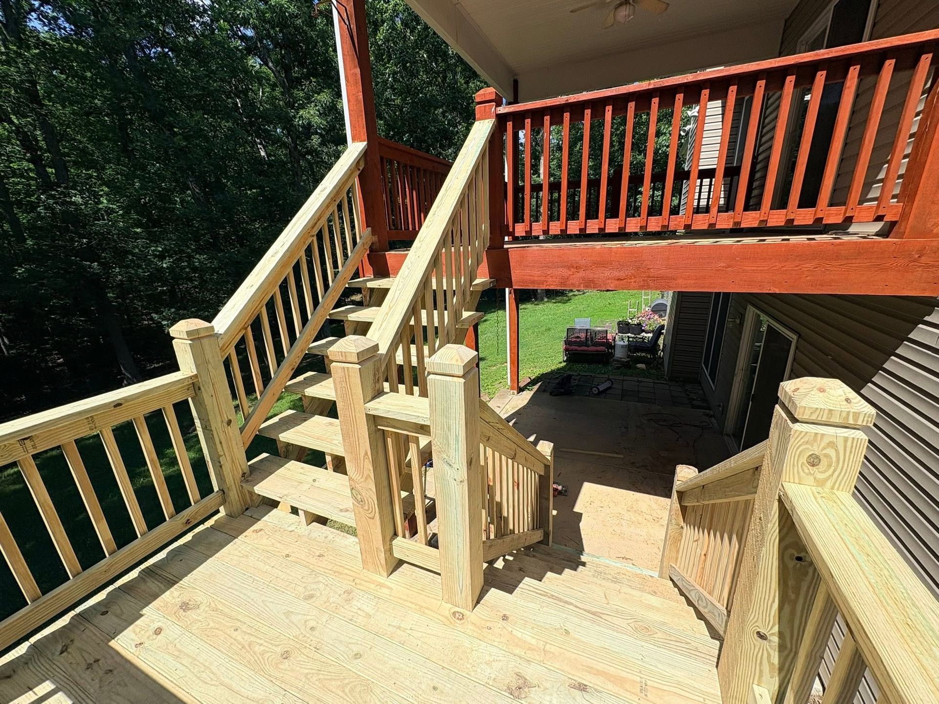 Wooden deck with stairs and railings leading to a raised porch beside a driveway and parked cars.