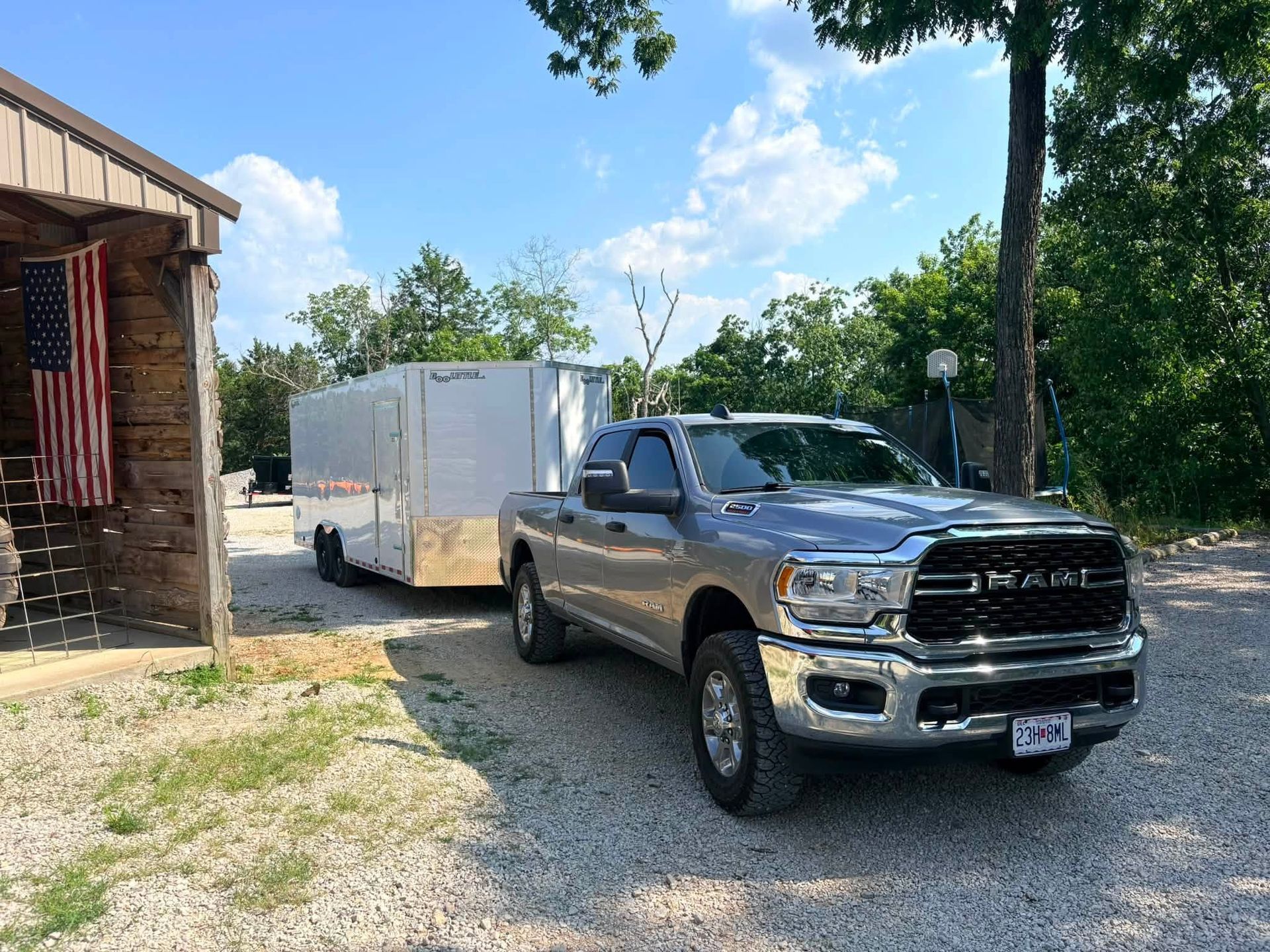 Silver pickup truck parked on a gravel driveway beside a white trailer near a wooden house.