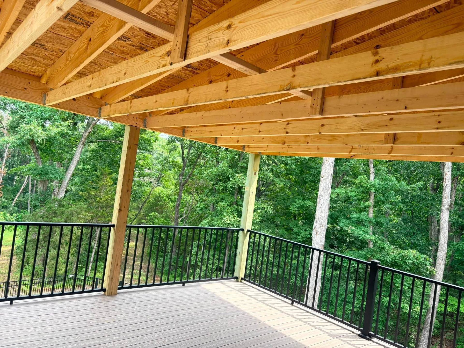 Covered wooden deck with black railing overlooking a green wooded area