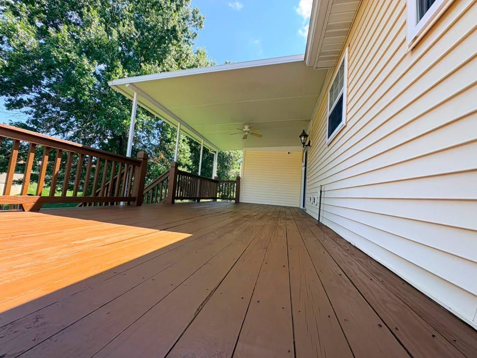 Back deck with tan siding, wooden railing, and awning beside a house on a sunny day
