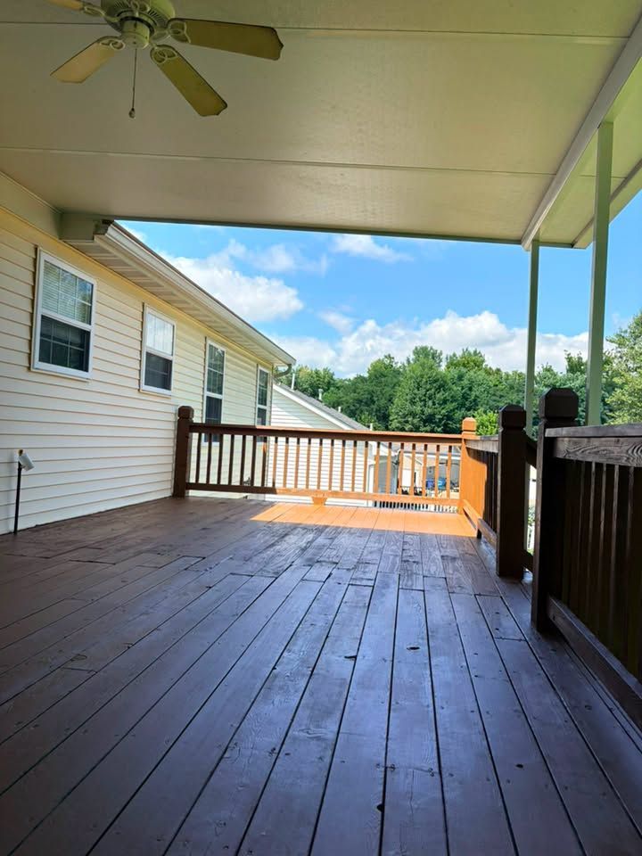 Covered wooden deck beside a house, with a ceiling fan, railing, and trees beyond.