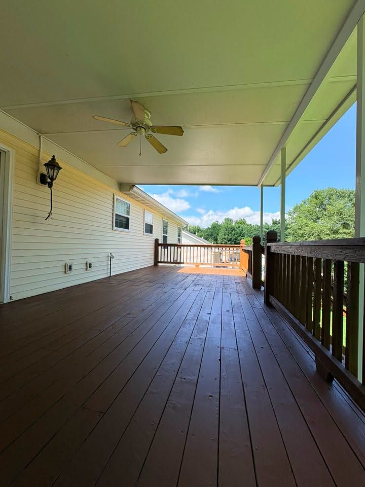 Covered wooden porch with brown floor, white ceiling fan, and black railing overlooking trees and sky