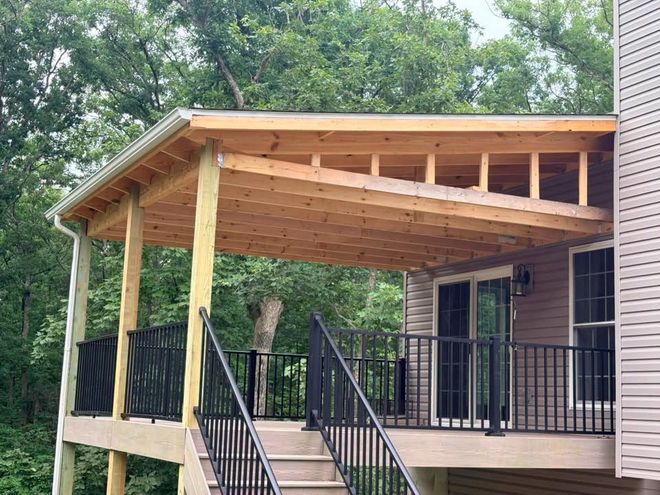 Covered wooden porch with black railings and stairs beside a brick house, surrounded by trees