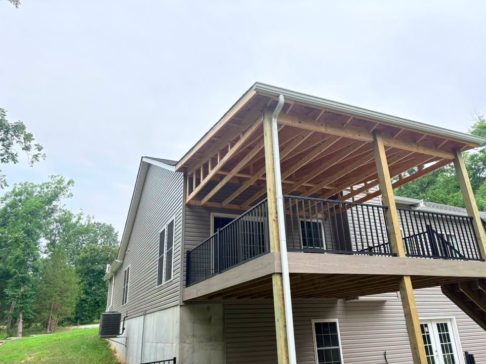 Raised beige house with covered deck and black railings, viewed from below under a cloudy sky