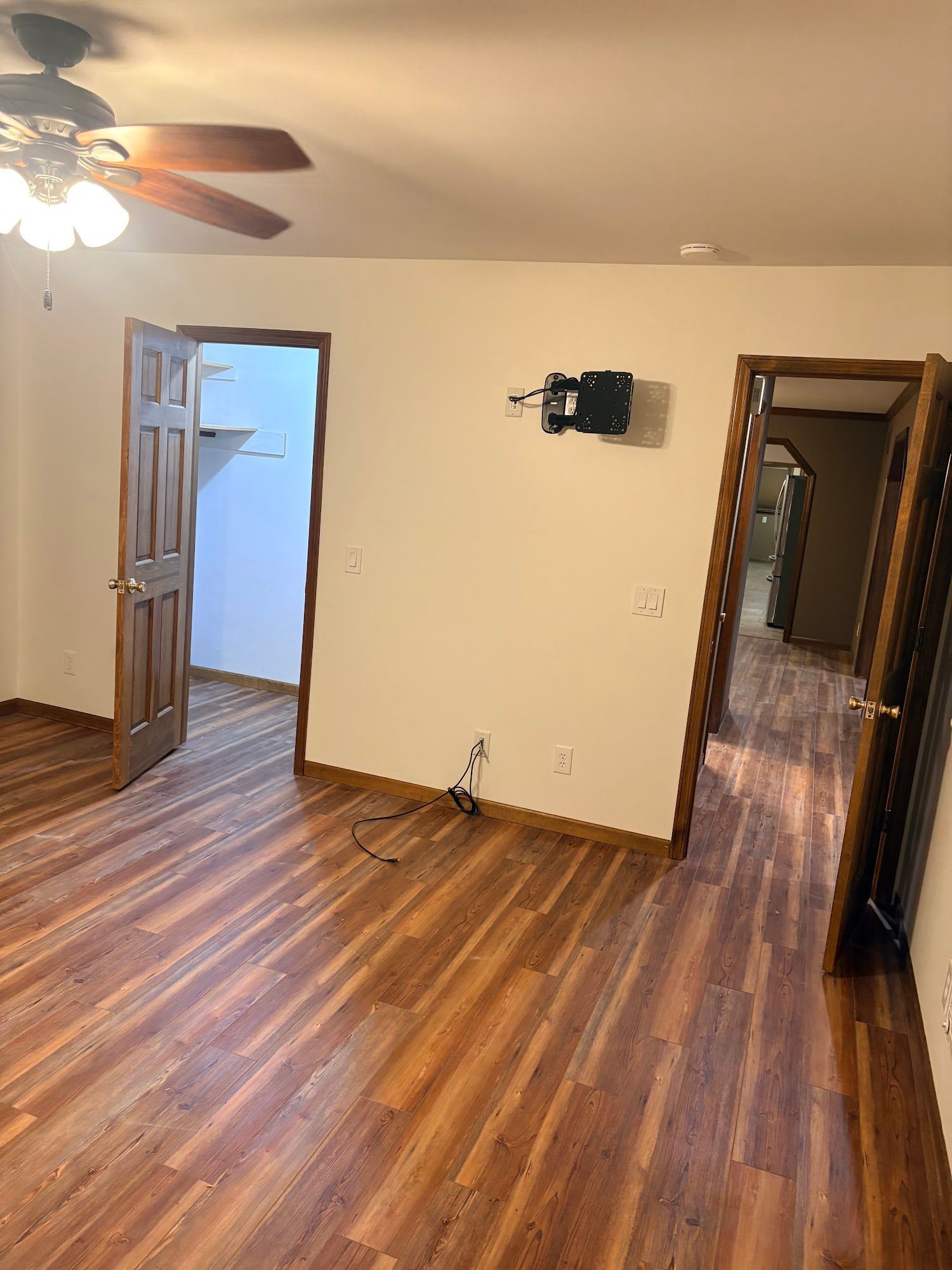Empty living room with wood floors, ceiling fan, open door, and wall-mounted TV bracket