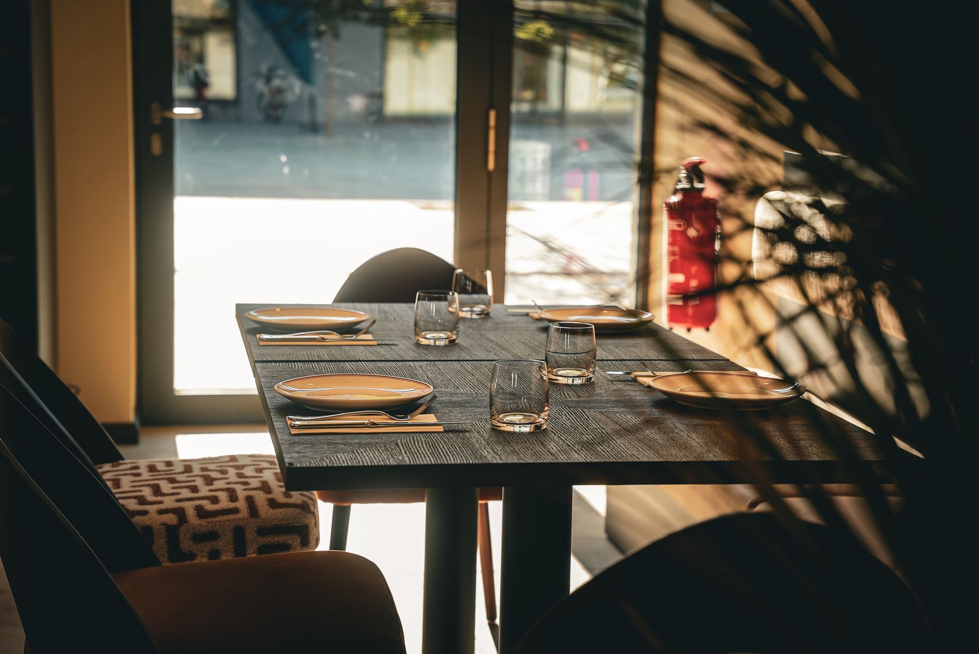 Ein sonnenbeschienener Café-Tisch mit Tellern und Gläsern neben einem Fenster und einem roten Feuerlöscher.