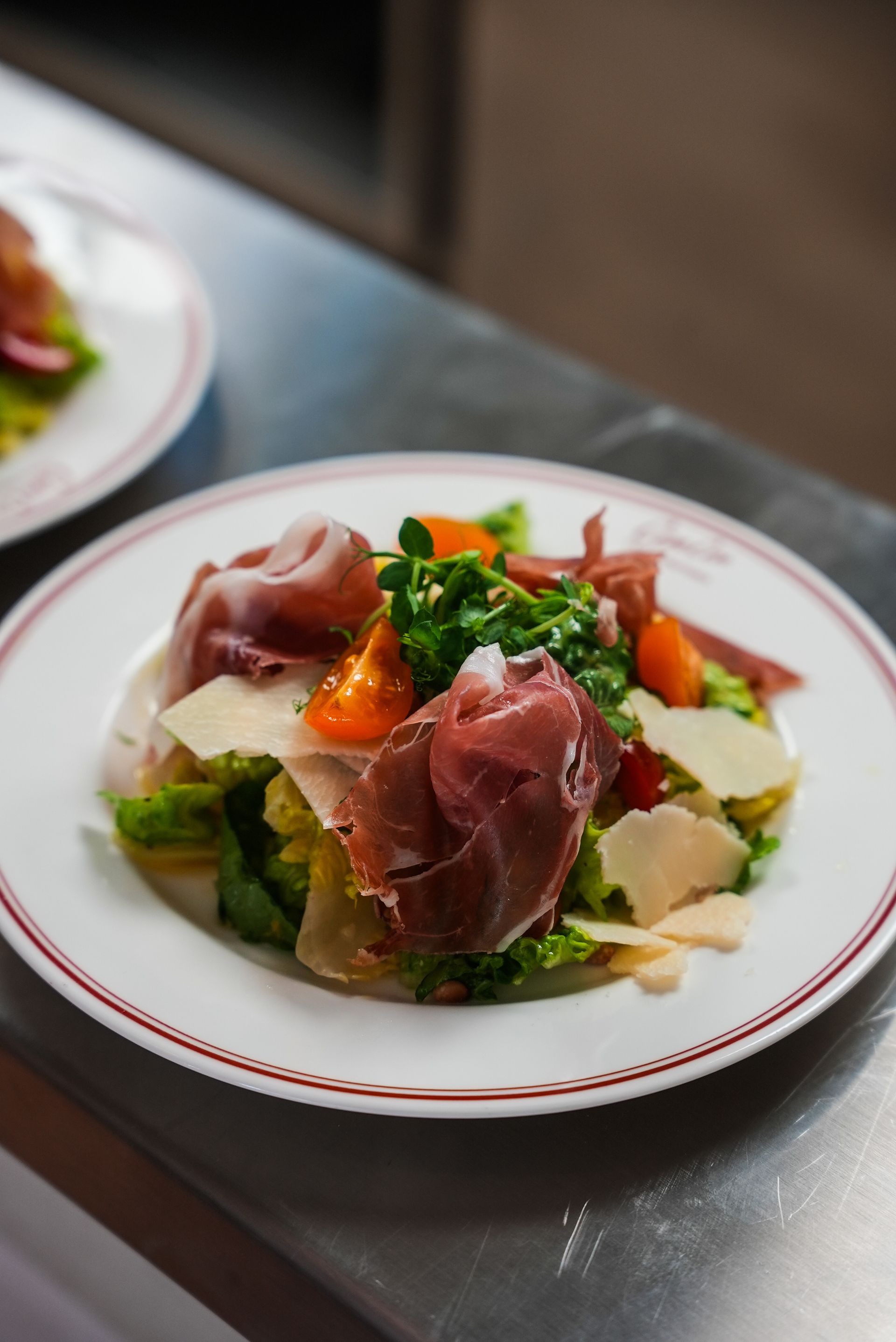Ein Teller Salat mit grünem Gemüse, Prosciutto, Tomaten und gehobeltem Käse auf einem Restauranttisch