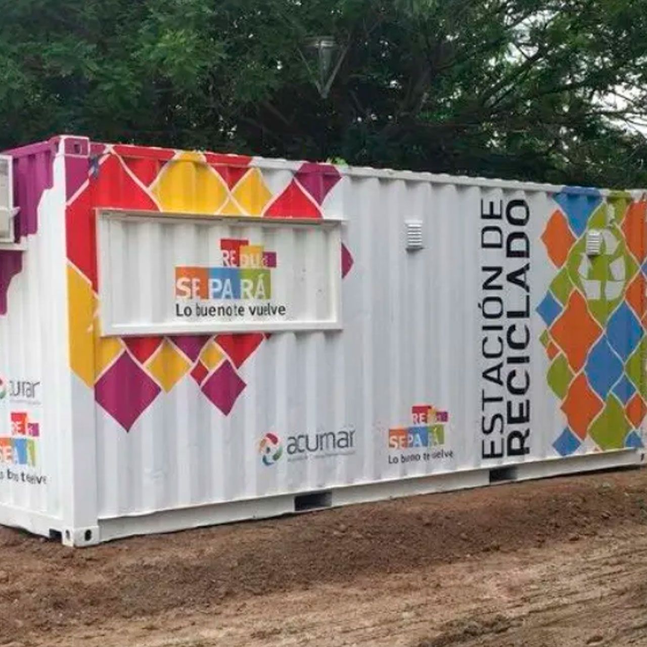 Contenedor de reciclaje blanco con coloridas pegatinas geométricas y letreros en español en una zona boscosa al aire libre.