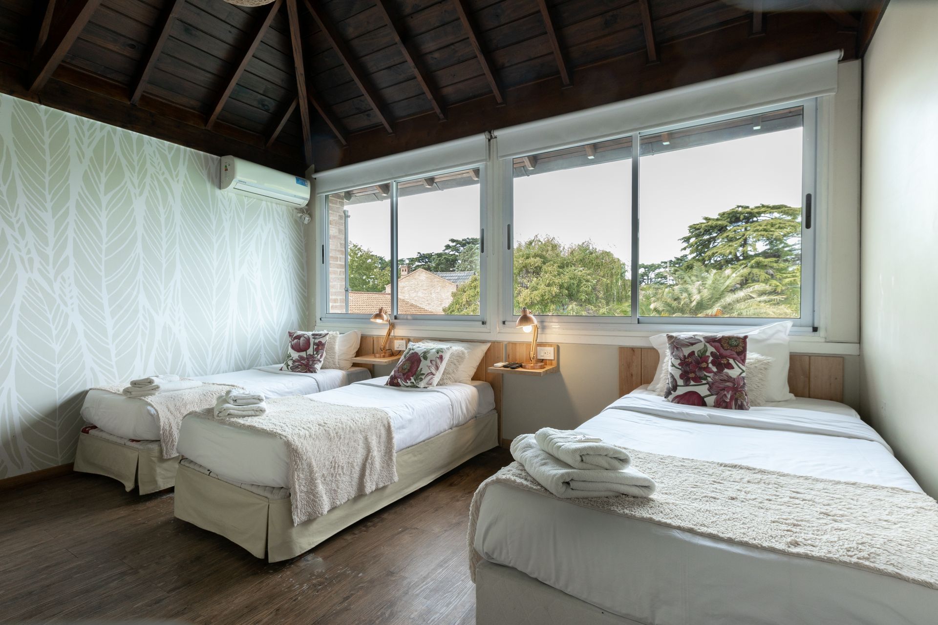 Acogedora habitación con tres camas blancas, techo de madera y grandes ventanales con vistas a la vegetación.
