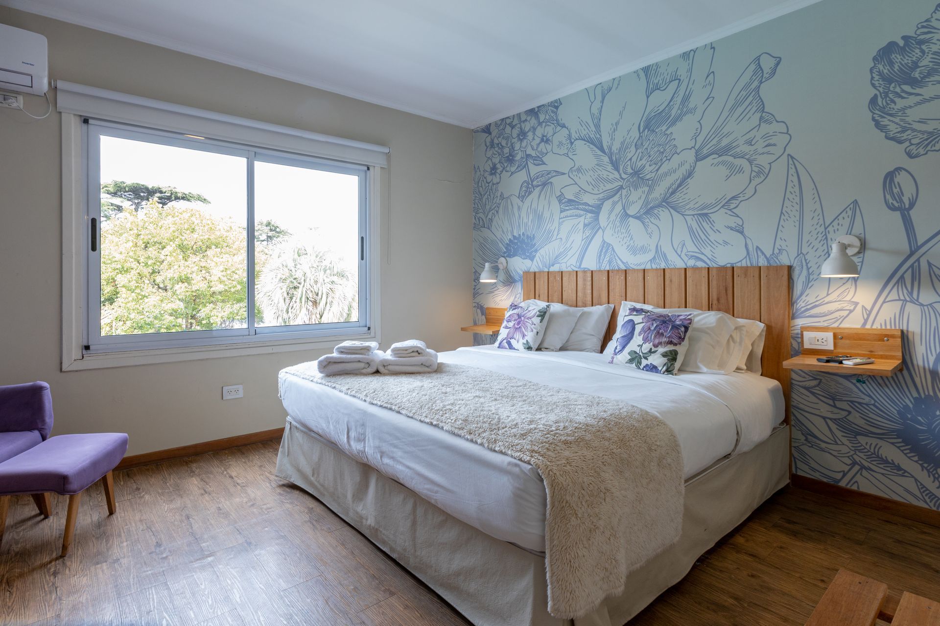 Habitación luminosa con una cama grande, un mural floral en la pared, luz natural en la ventana y un puf morado.