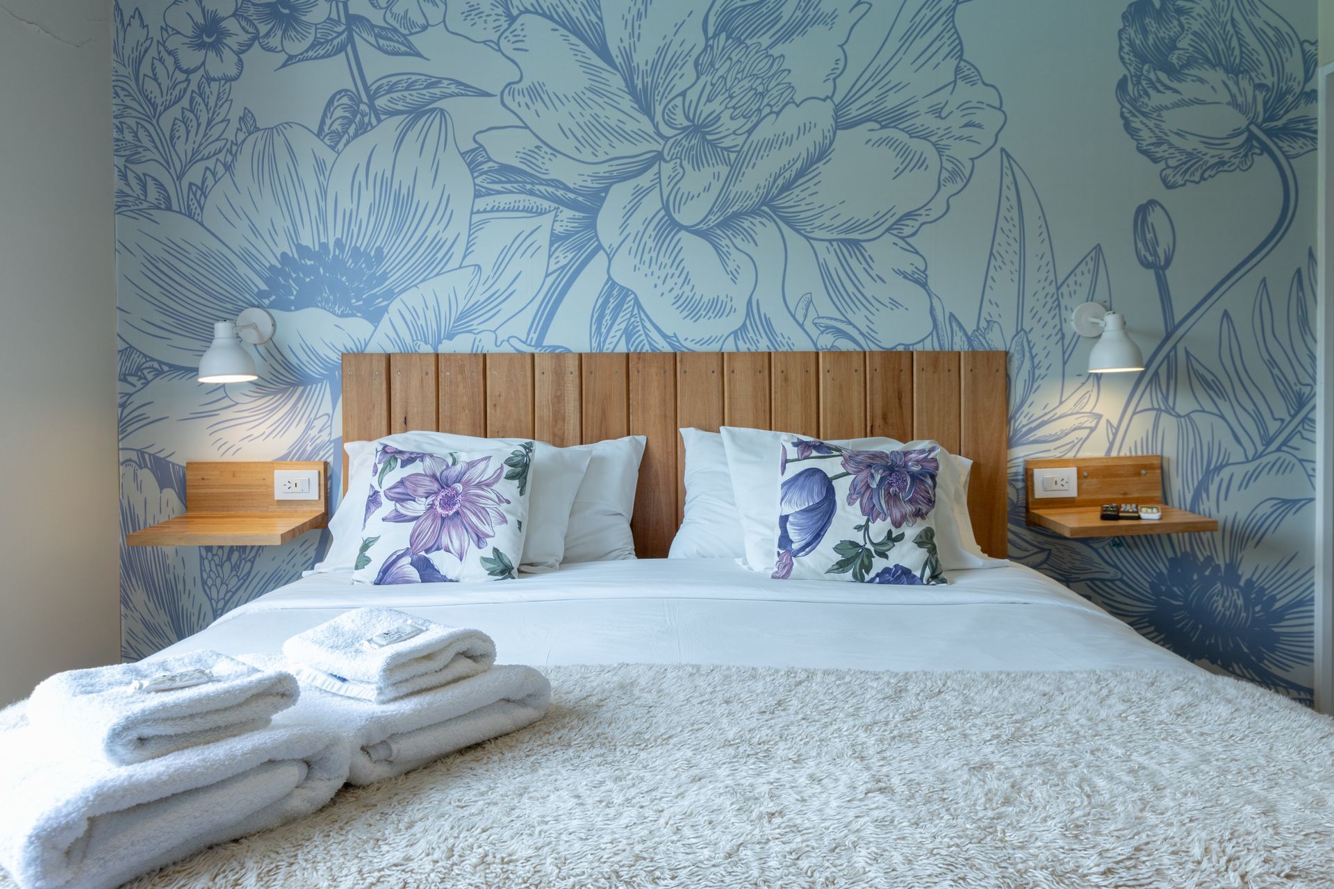 Cama bien hecha con papel pintado floral, almohadas azules, lámparas de noche y toallas dobladas a los pies.